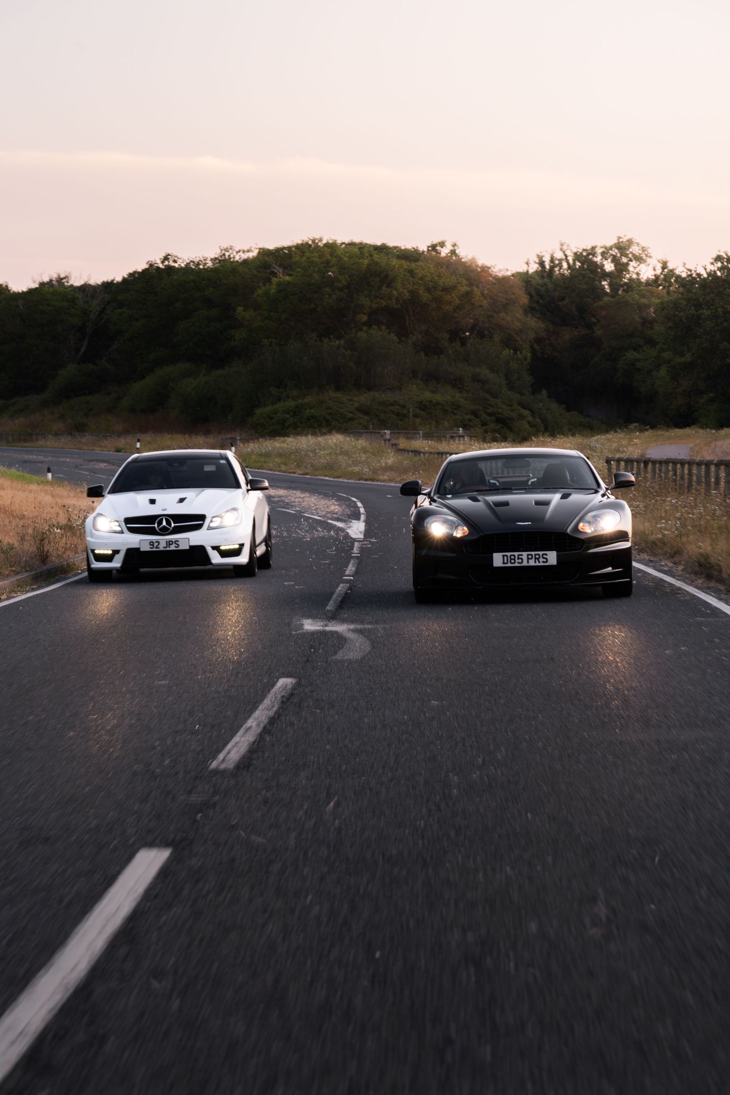 White Mercedes and black Aston Martin driving side by side on an open road at dusk.