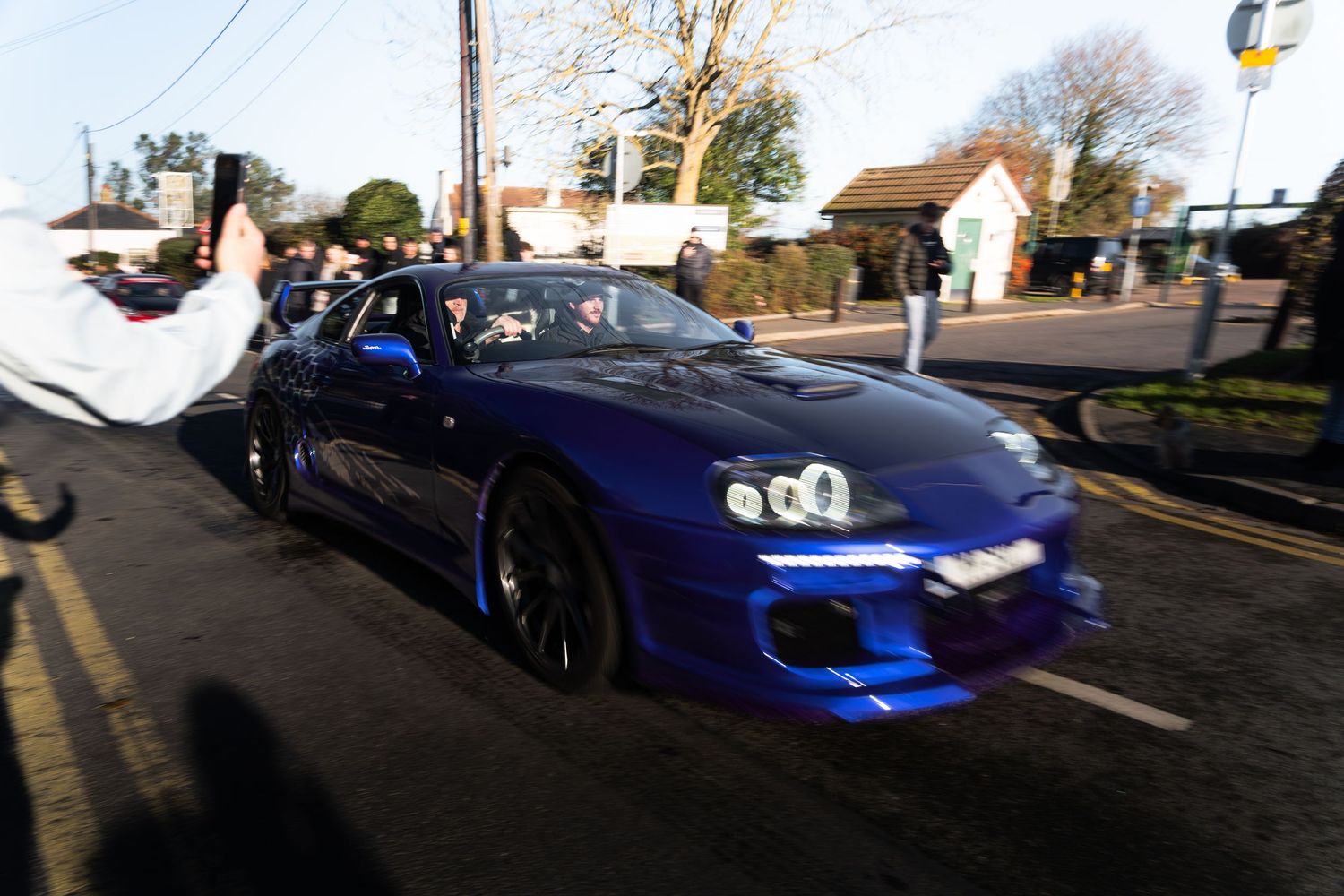 A modified blue Toyota Supra sports car driving on a UK street with spectators watching.
