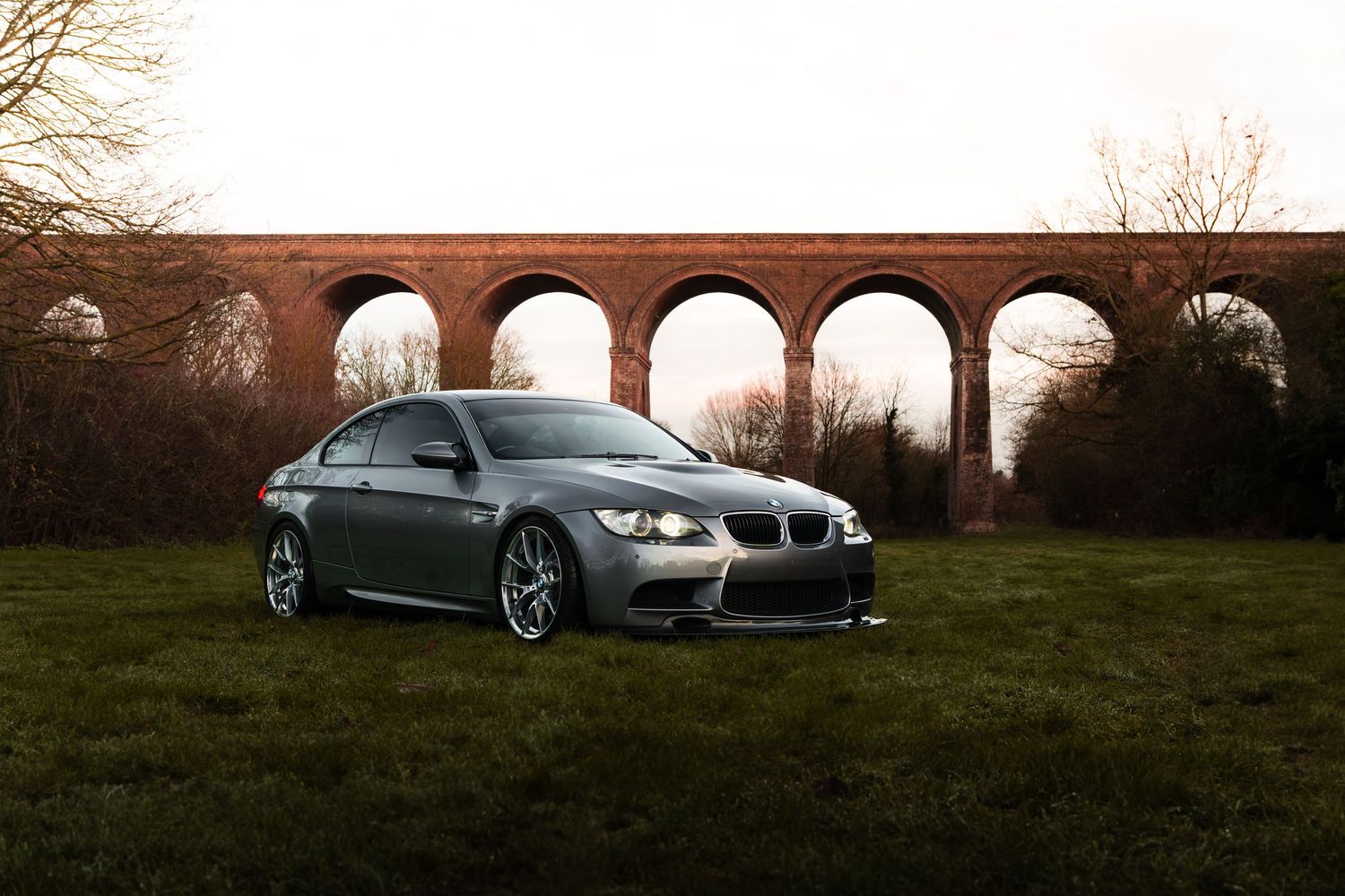 A silver BMW M3 coupe parked on grass in front of a large brick railway viaduct.