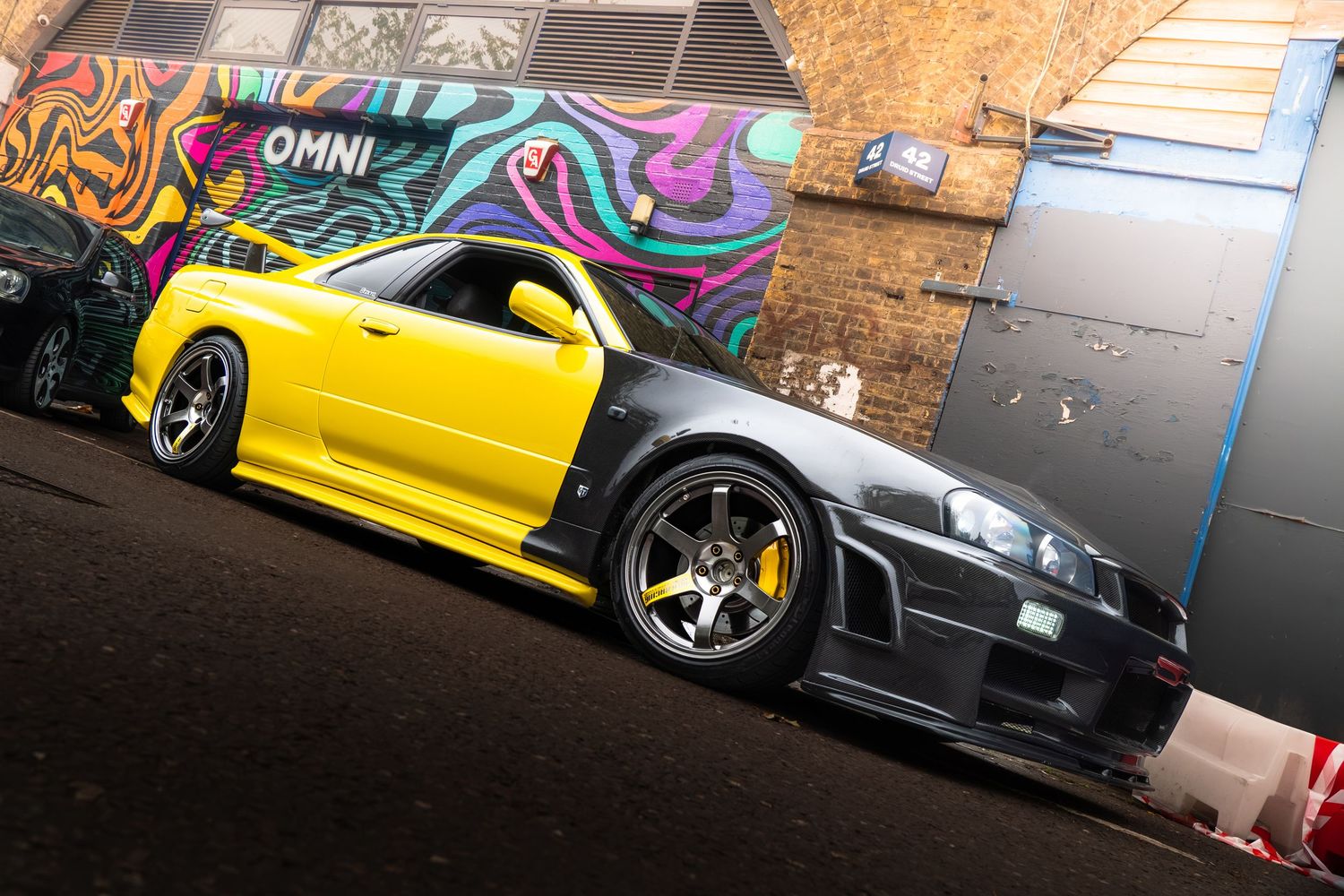 A modified yellow and black Nissan Skyline R34 GT-R parked in front of a colorful graffiti wall.