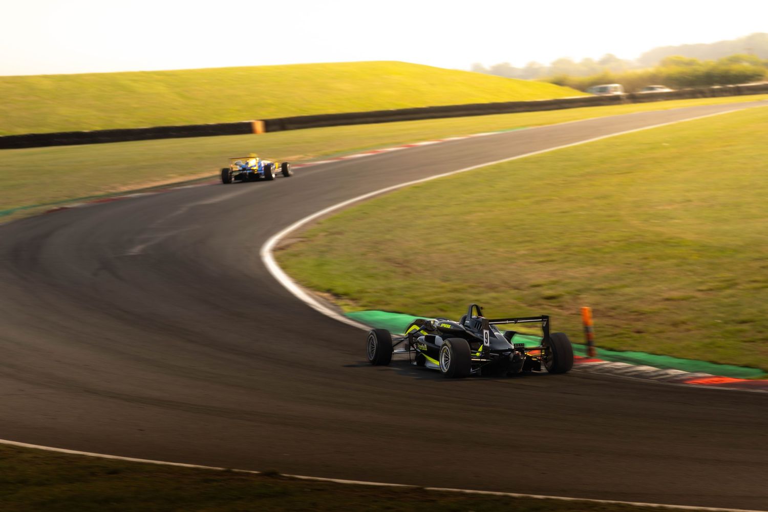 Black Formula racing car leads through a sweeping bend on a scenic sunlit race circuit.