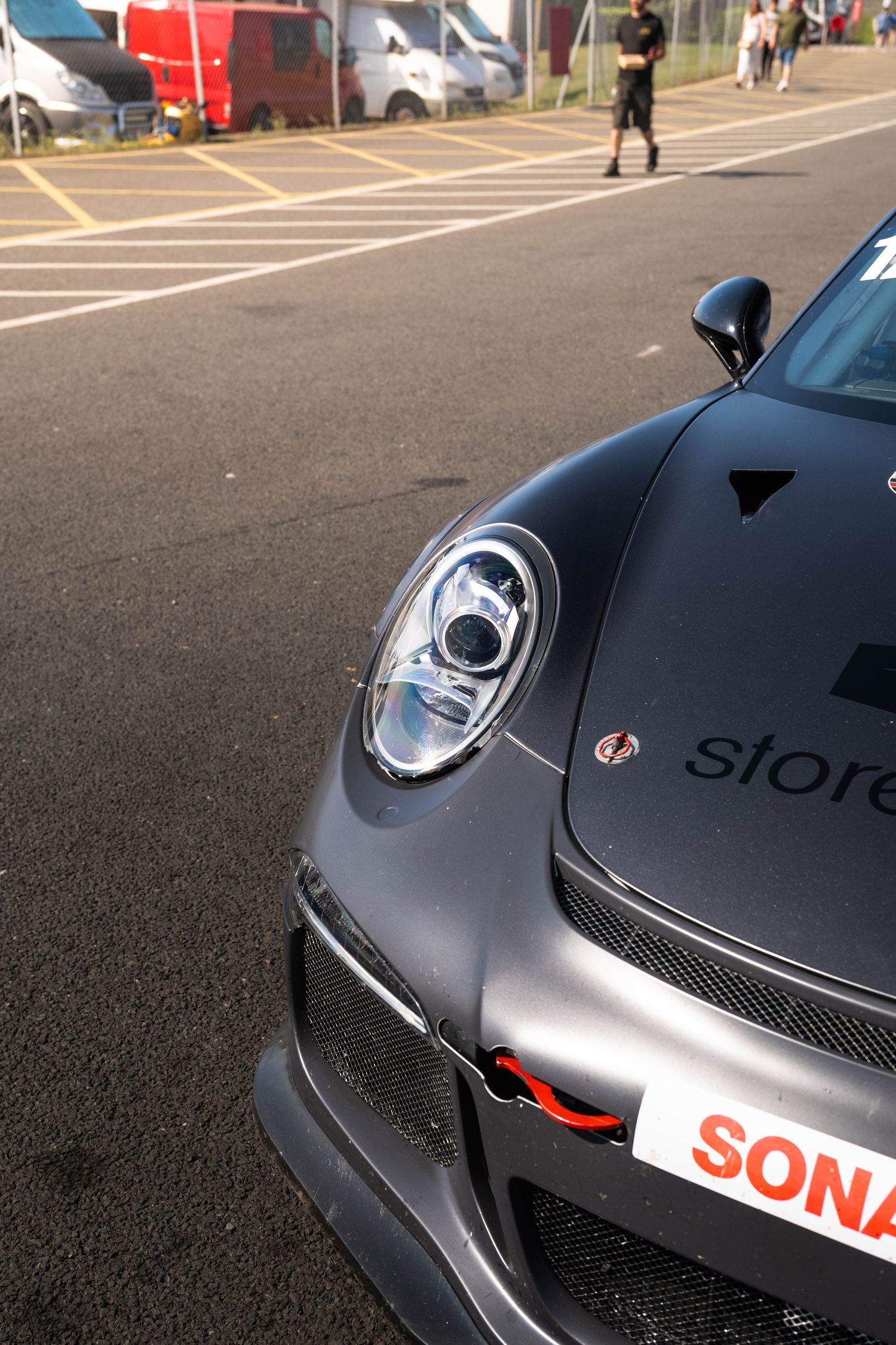 Close-up of a dark grey Porsche 911 GT3 front headlight and bumper with SONAX branding in paddock.