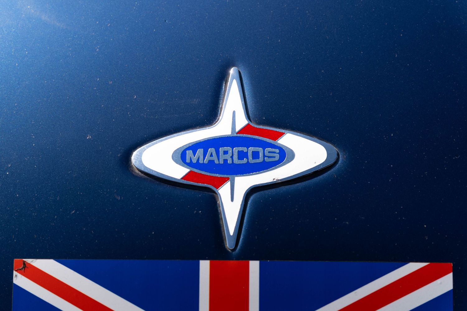 Marcos car badge emblem in chrome and blue with Union Jack flag reflection on a dark blue hood.
