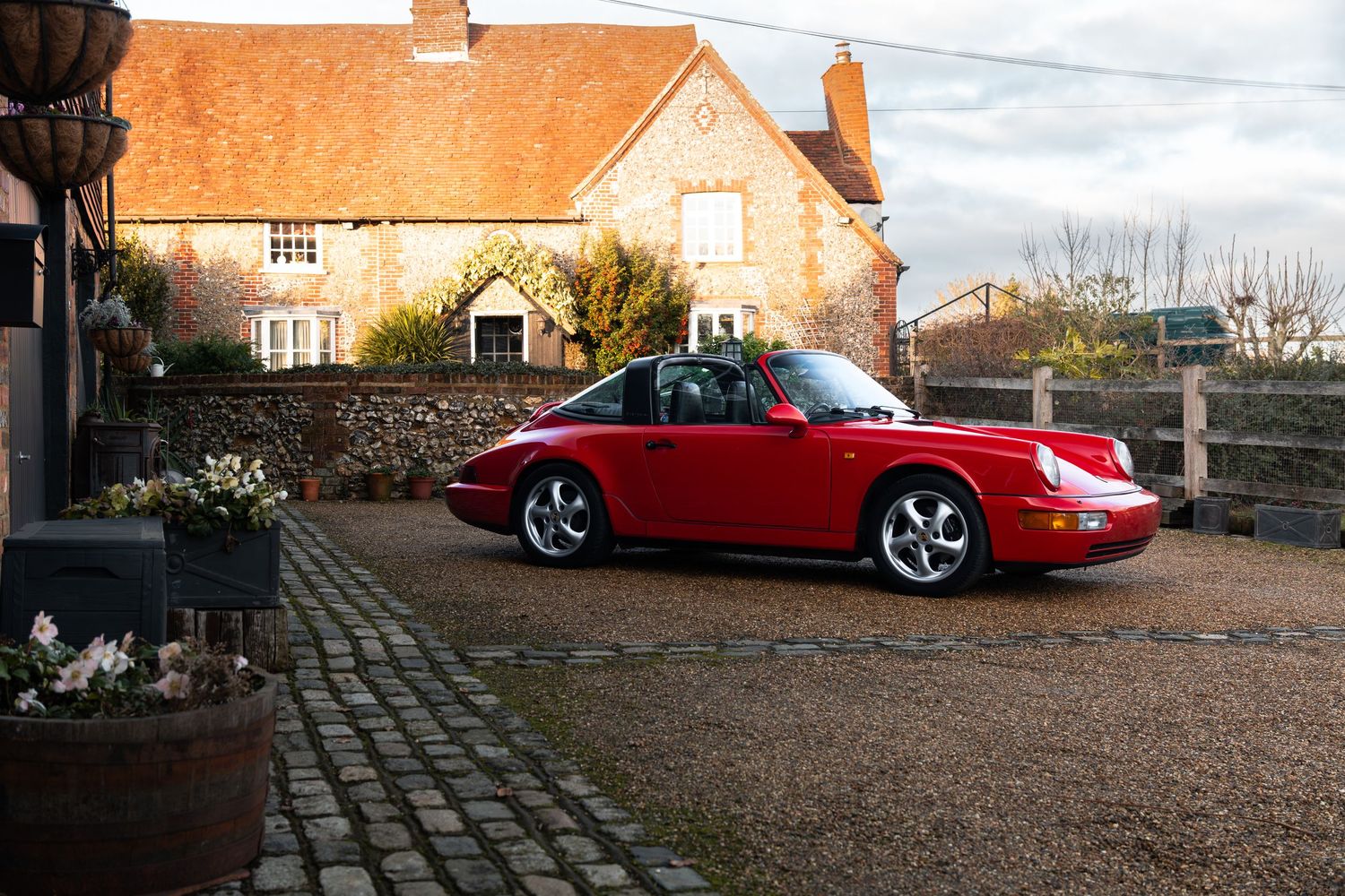 Red Porsche 911 Carrera parked on cobblestone driveway beside a rustic English countryside stone cottage.