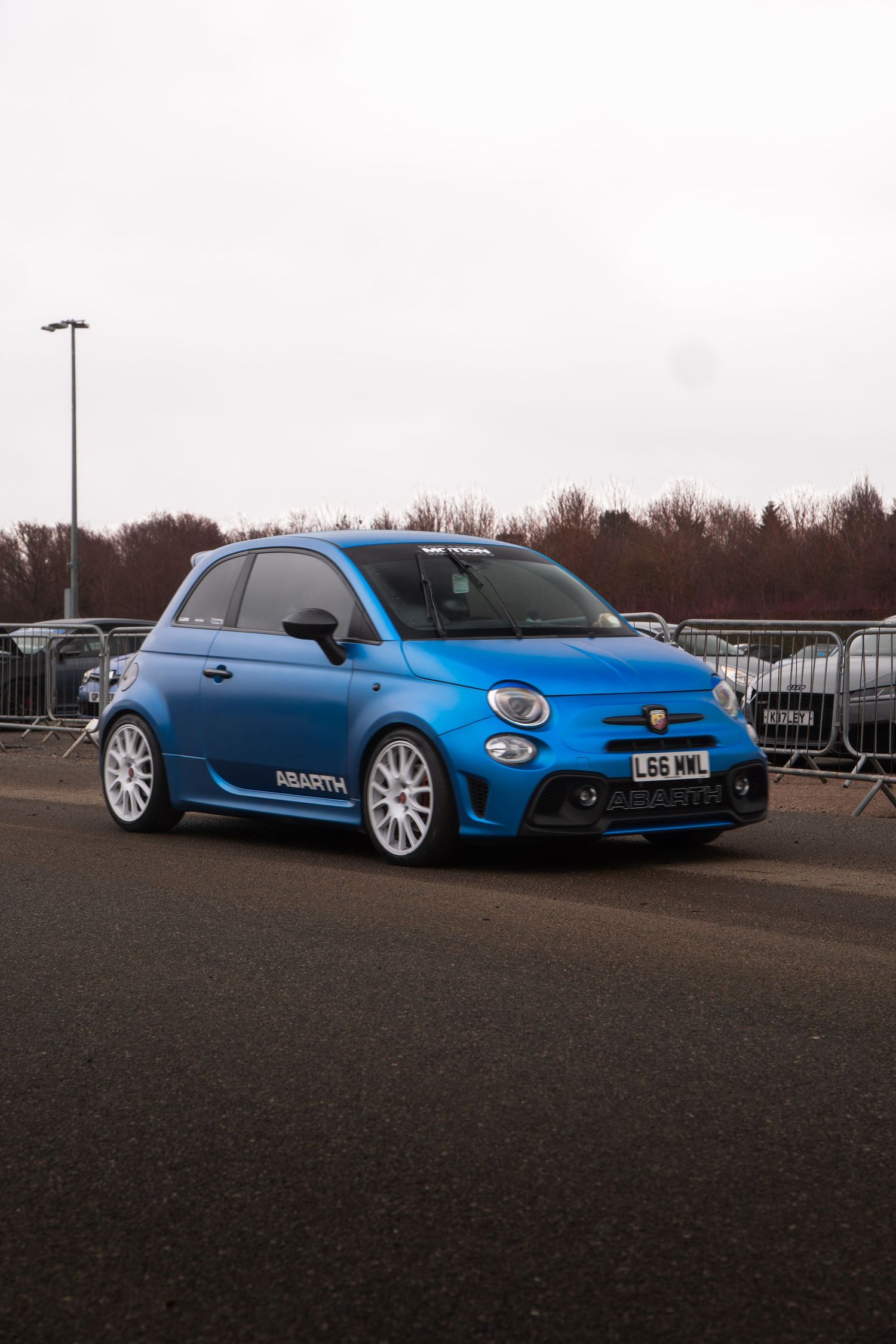 Blue Abarth 595 hatchback with white alloys parked at a motorsport event.