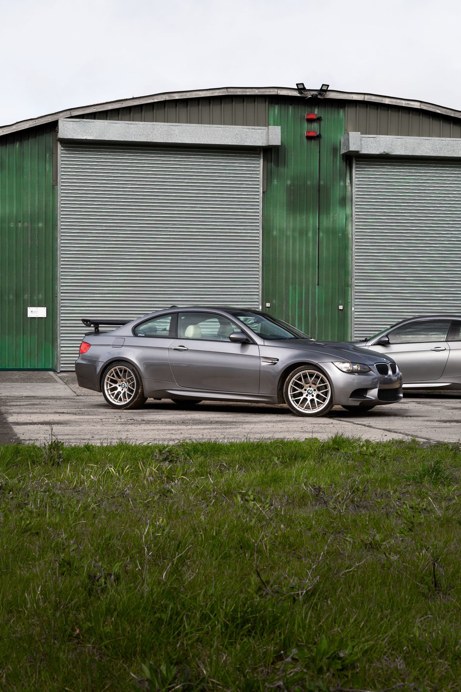 Gray BMW M3 coupe with rear spoiler parked in front of green industrial garage doors.