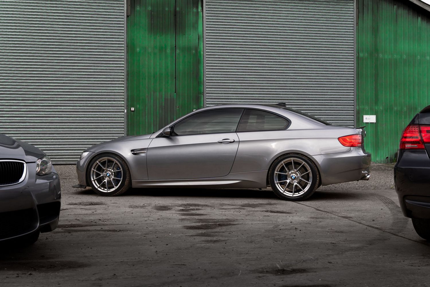 Side profile of a lowered silver BMW M3 coupe parked against corrugated metal walls.