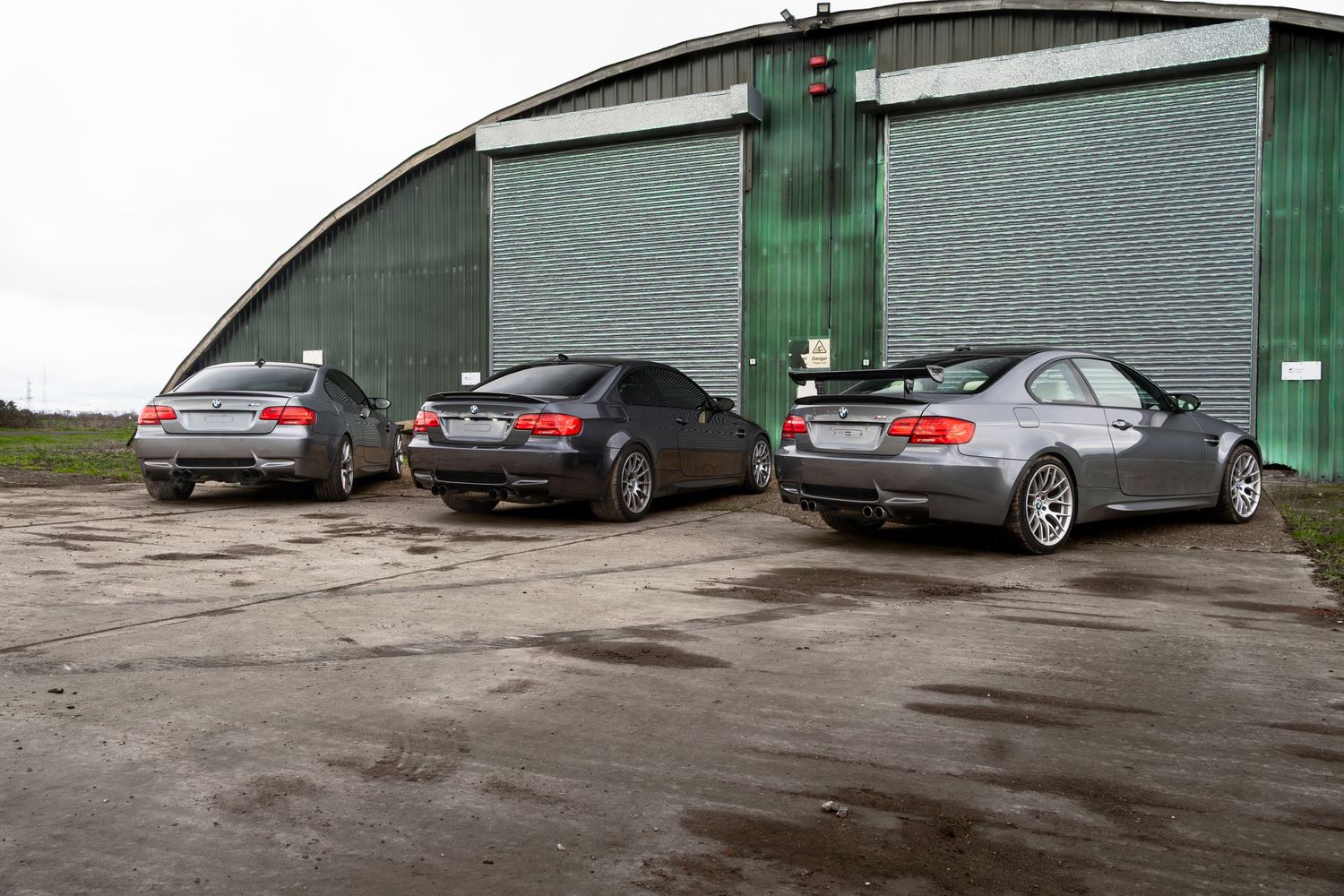 Three BMW M3 coupes parked outside a large green arched metal warehouse building.