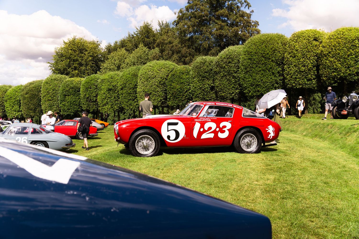 Vintage red Ferrari race car number 5 on display at an outdoor classic car show on green grass.