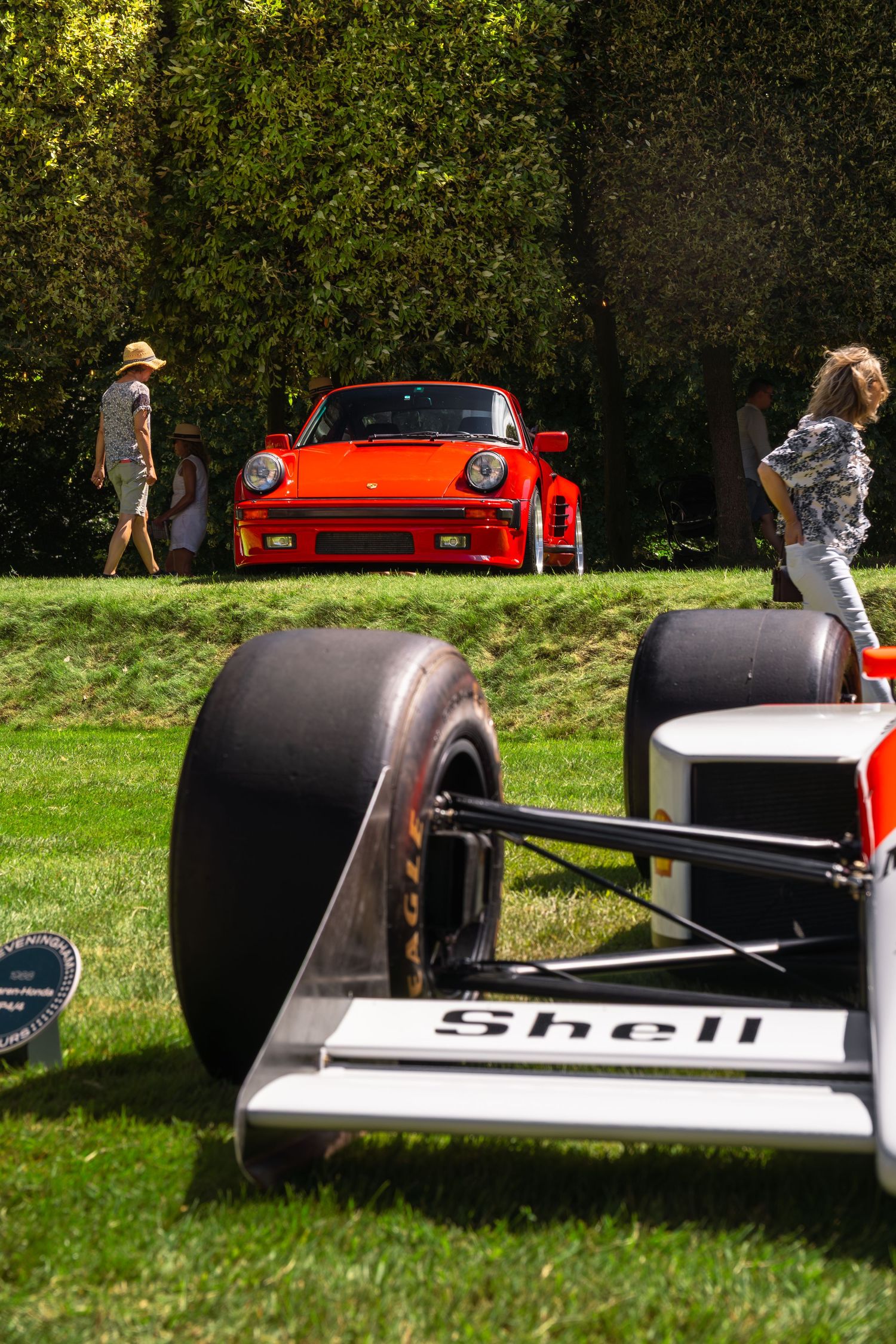 A red Porsche 911 on grass at a car show, with a Shell-sponsored Formula 1 car in the foreground.