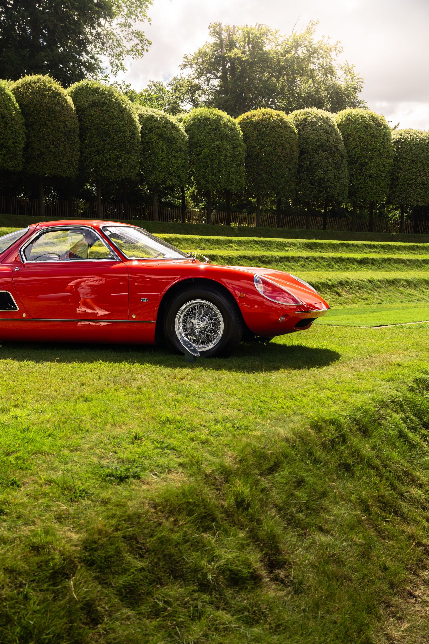 Vintage red Ferrari sports car parked on lush green grass with manicured hedges and trees in background.
