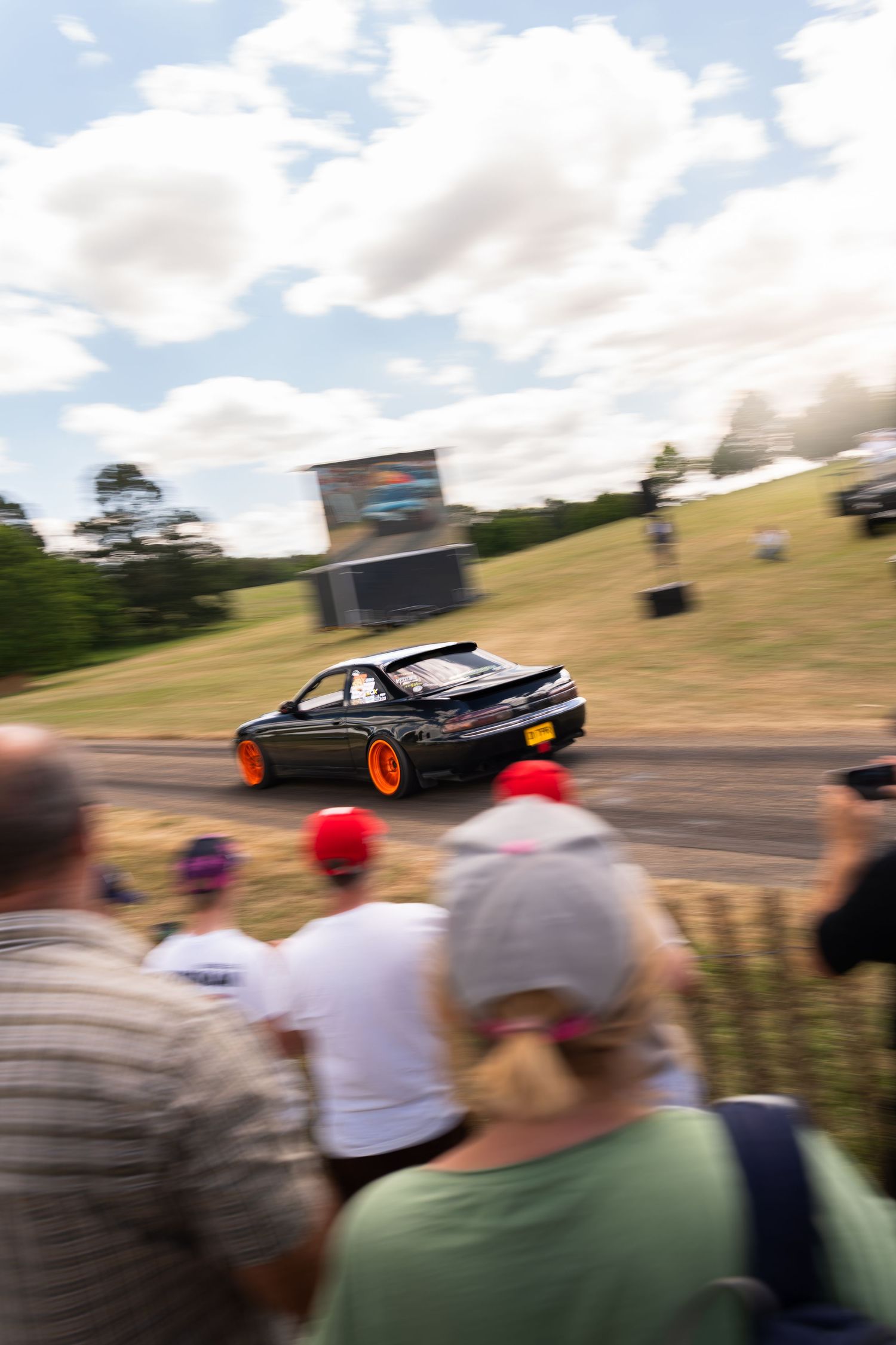 Black car with orange wheels drifting on a race track while spectators watch at an outdoor motorsport event.