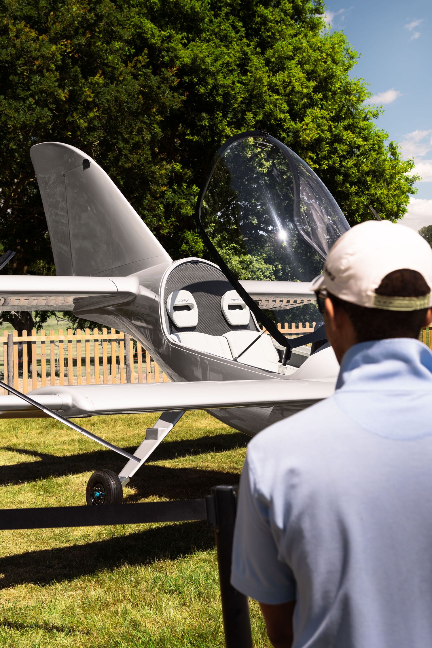 Man in white cap examines a small light aircraft on grass at an outdoor airshow on a sunny day.