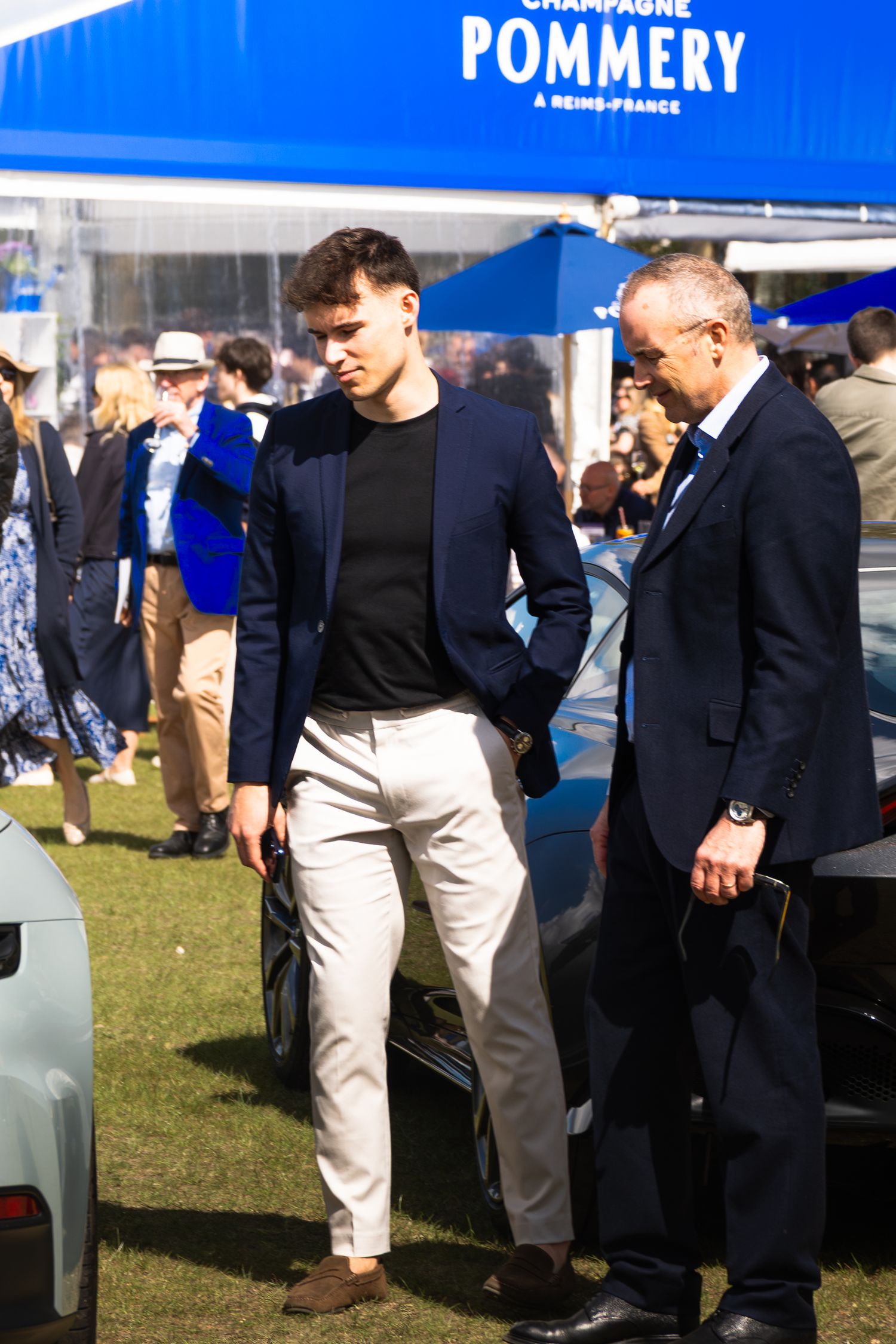Two well-dressed men in navy blazers at a Pommery Champagne luxury automotive event outdoors.