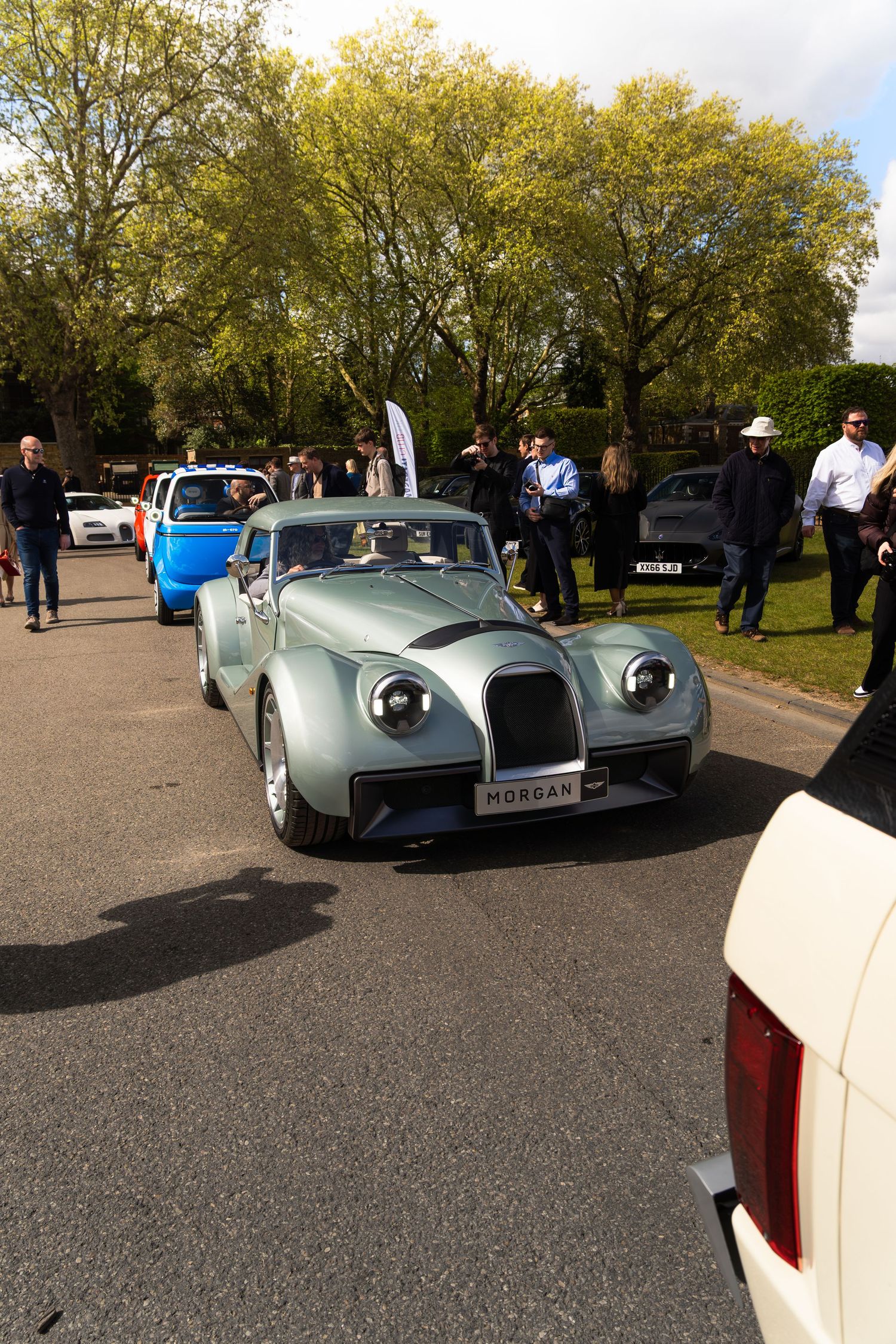 A vintage silver Morgan sports car on display at an outdoor classic car show surrounded by spectators.