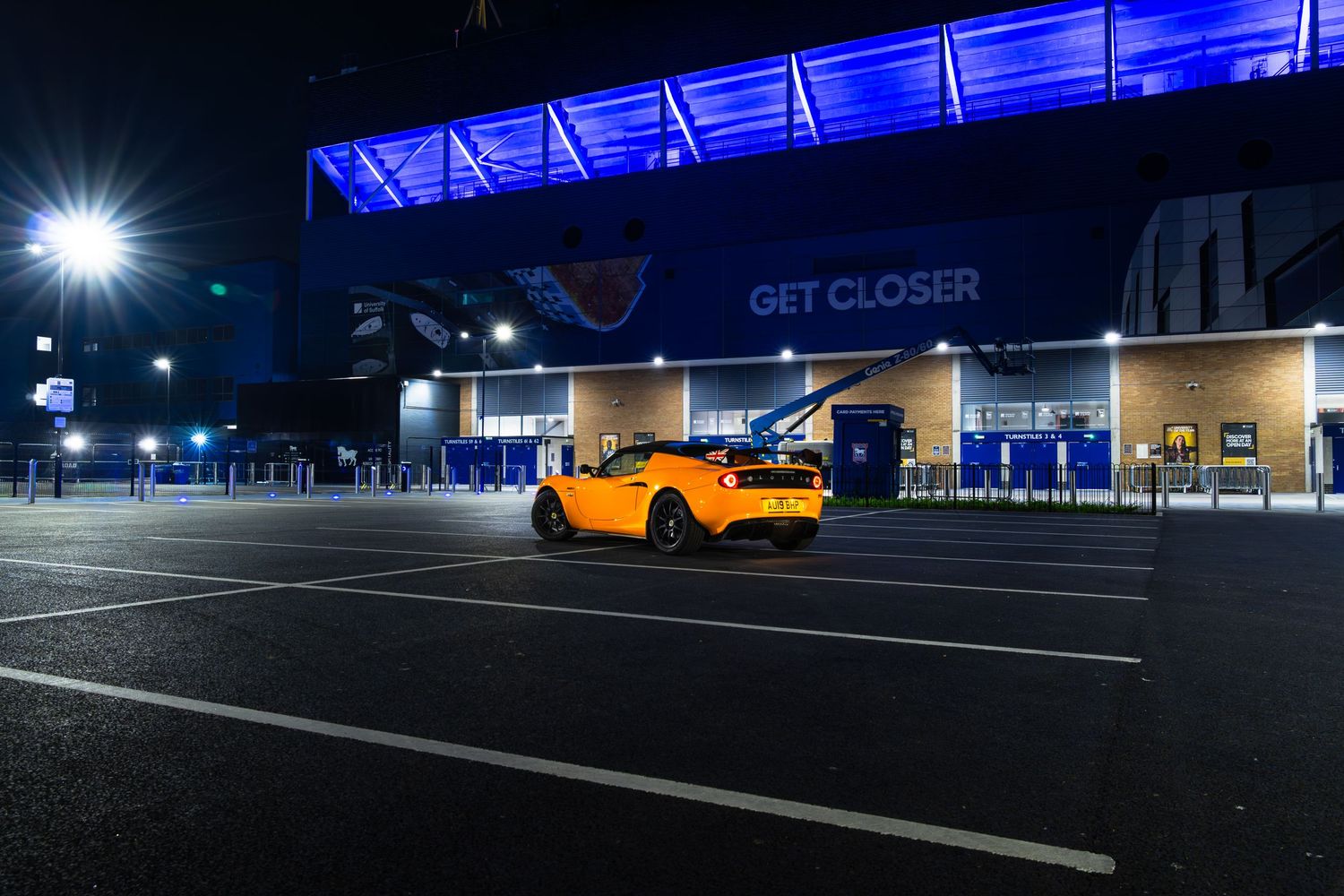 Orange Lotus Elise sports car parked in empty lot outside blue-lit GET CLOSER football stadium at night.
