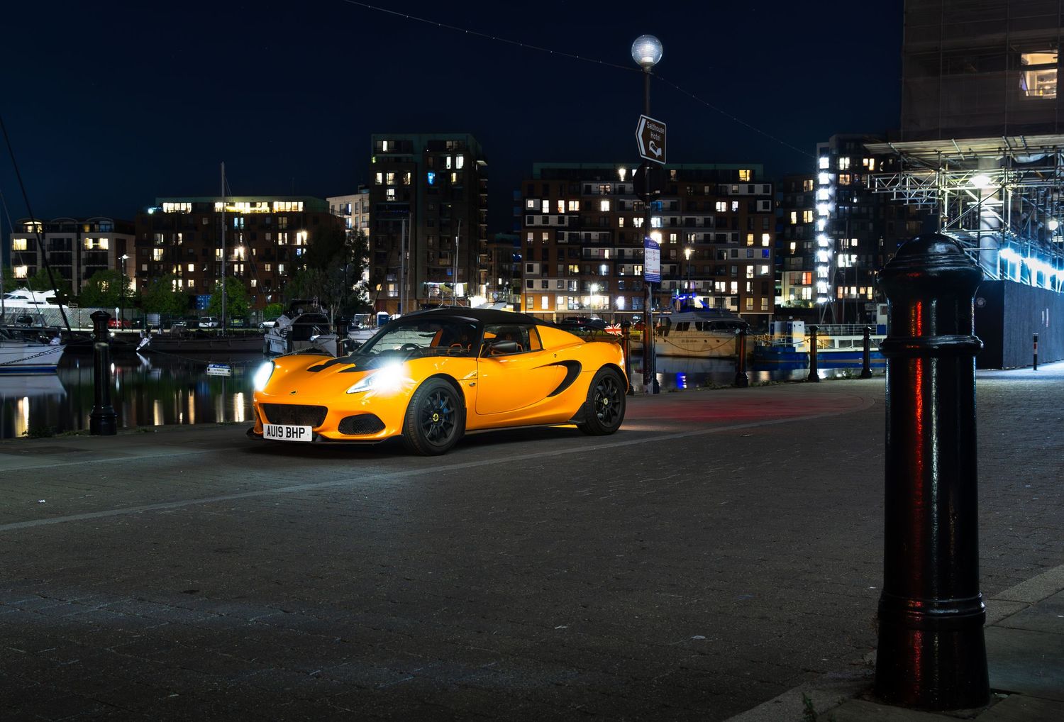 Yellow Lotus Elise sports car photographed at night near a marina with glowing city buildings.