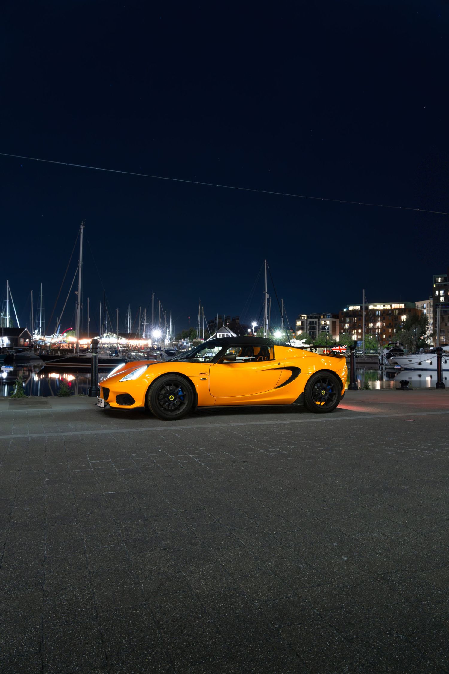 Yellow Lotus Elise sports car parked at a marina at night with sailboats in the background.