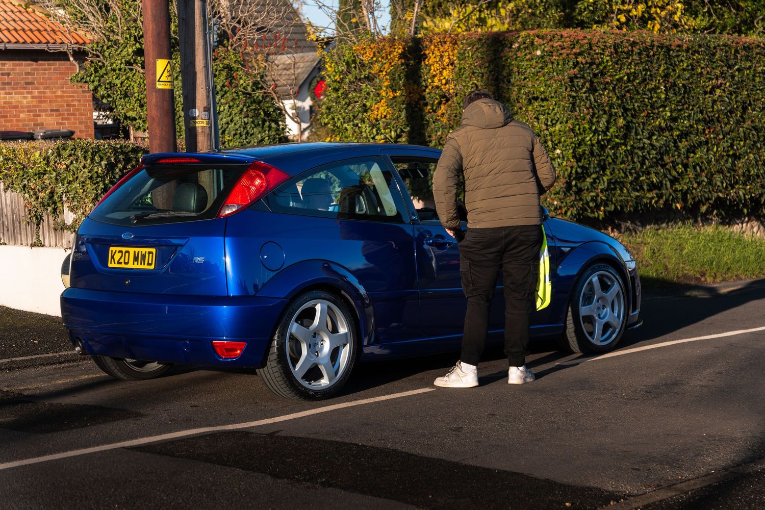 Person in hoodie standing beside a blue Ford Focus RS hatchback parked on a sunny suburban street.