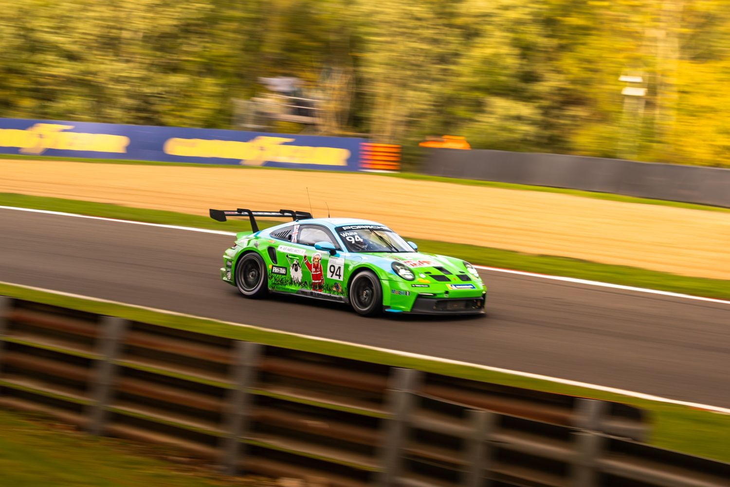 Bright green Porsche GT3 race car speeding along circuit with blurred autumn foliage background.