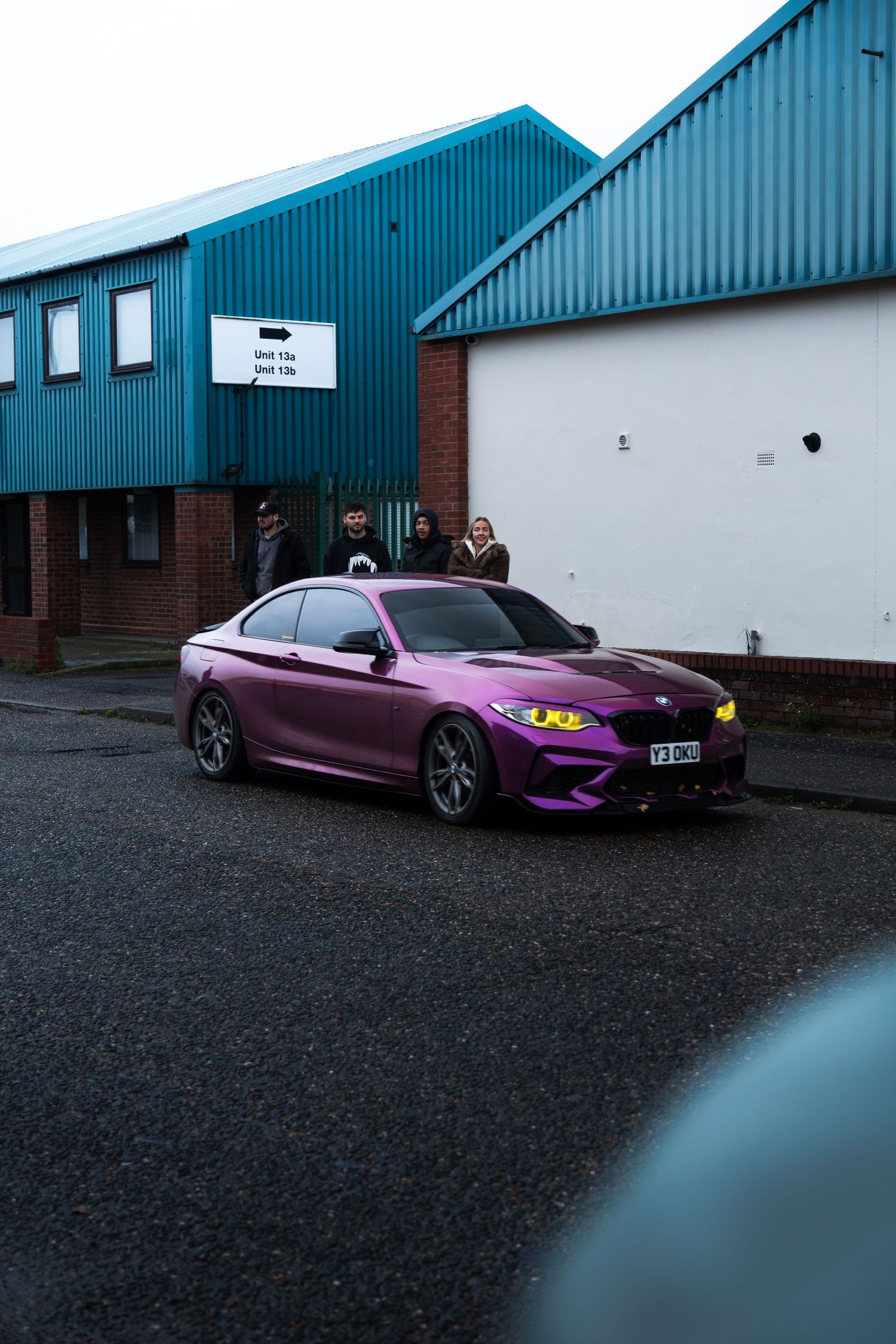 A purple wrapped BMW M2 with yellow headlights parked outside an industrial building.
