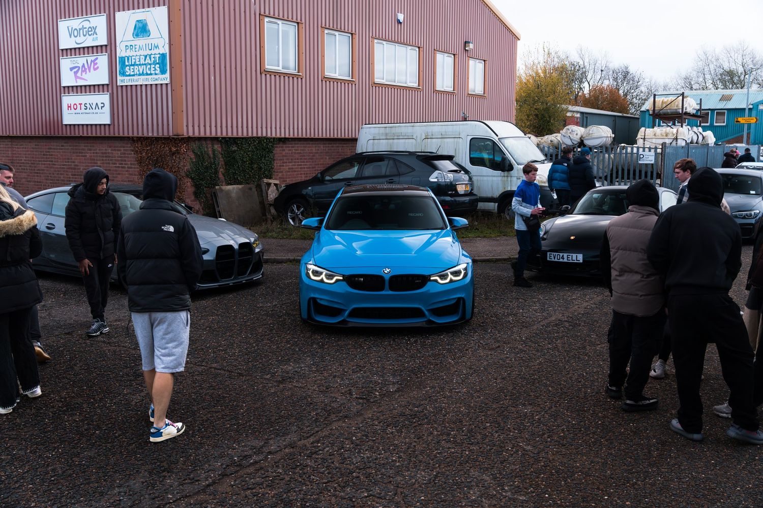 A blue BMW M4 surrounded by car enthusiasts at an outdoor automotive meet event.