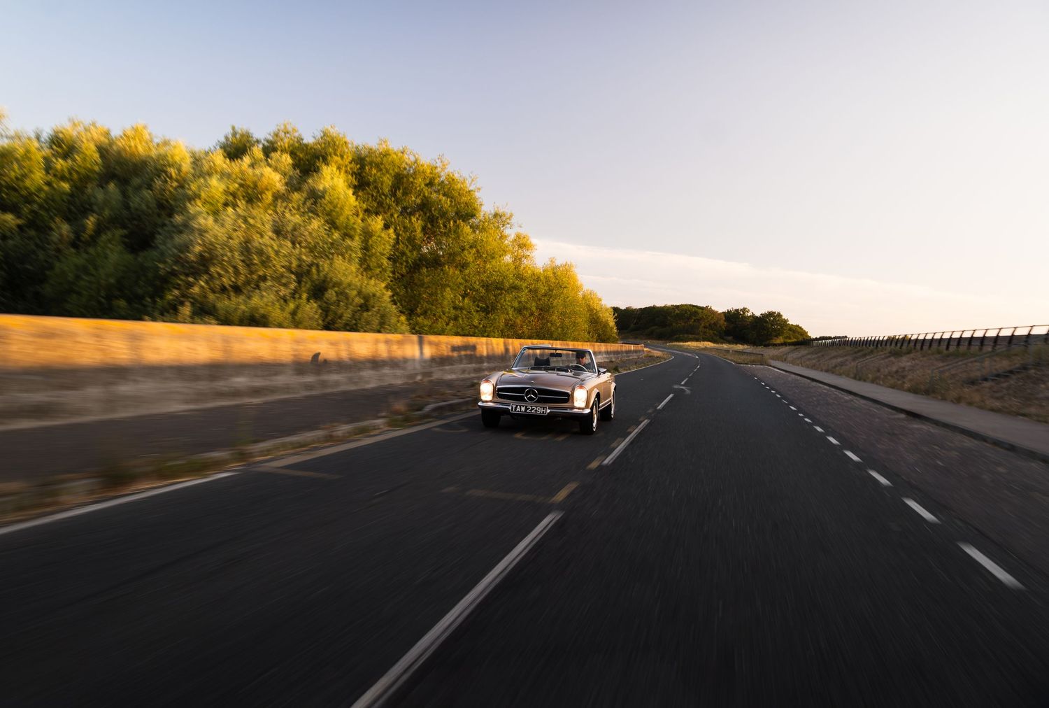 A classic white convertible car driving on an open highway at golden hour with lush green trees in the background.