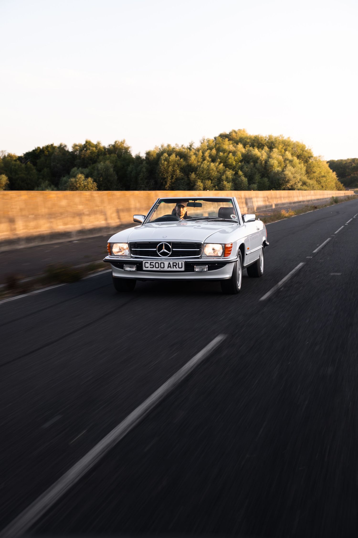 White Mercedes-Benz SL convertible driving on open road at golden hour, UK registration plate, trees in background.