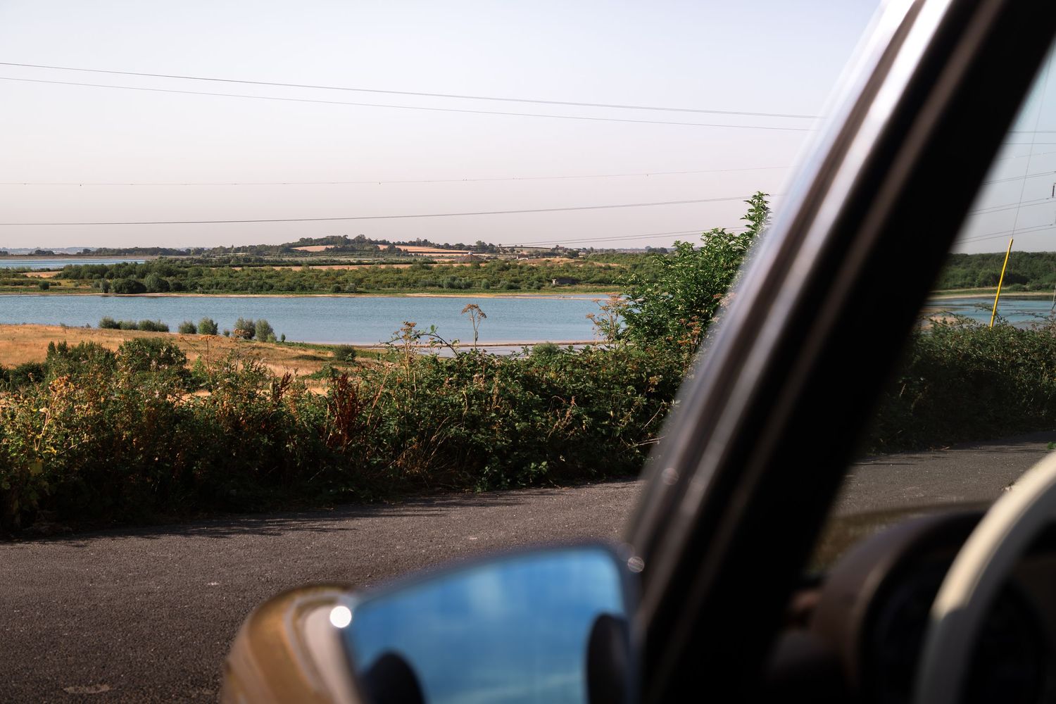 View from a car window of a serene lake surrounded by green shrubs and fields under a clear evening sky.