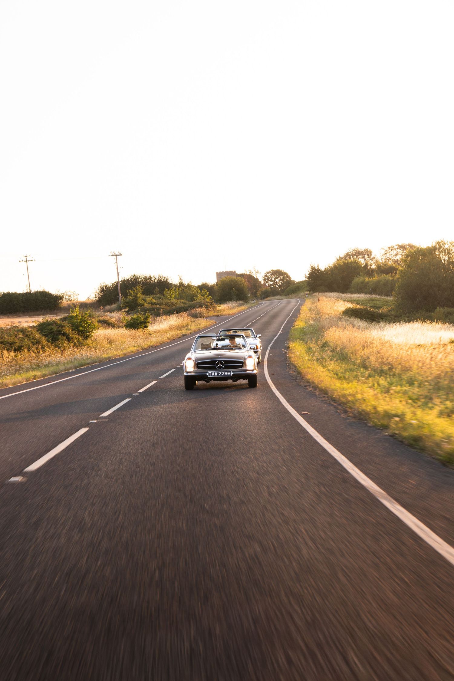 A classic convertible car driving on a straight rural road at golden hour sunset.