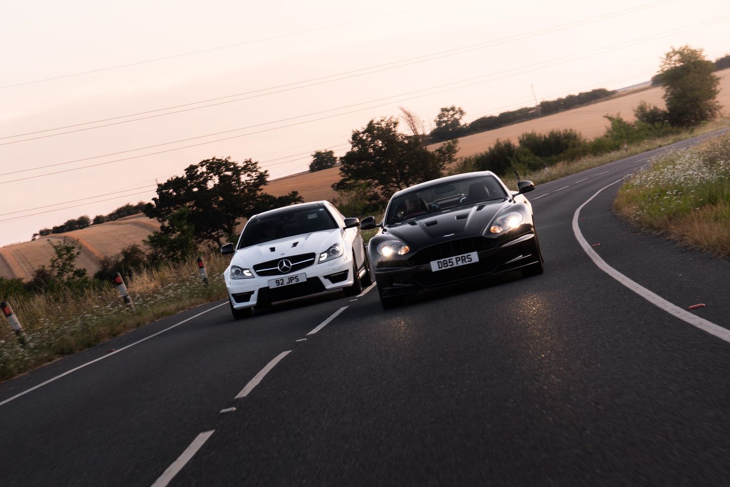 Two sports cars, a white Mercedes and dark Aston Martin, racing on a rural road at dusk.
