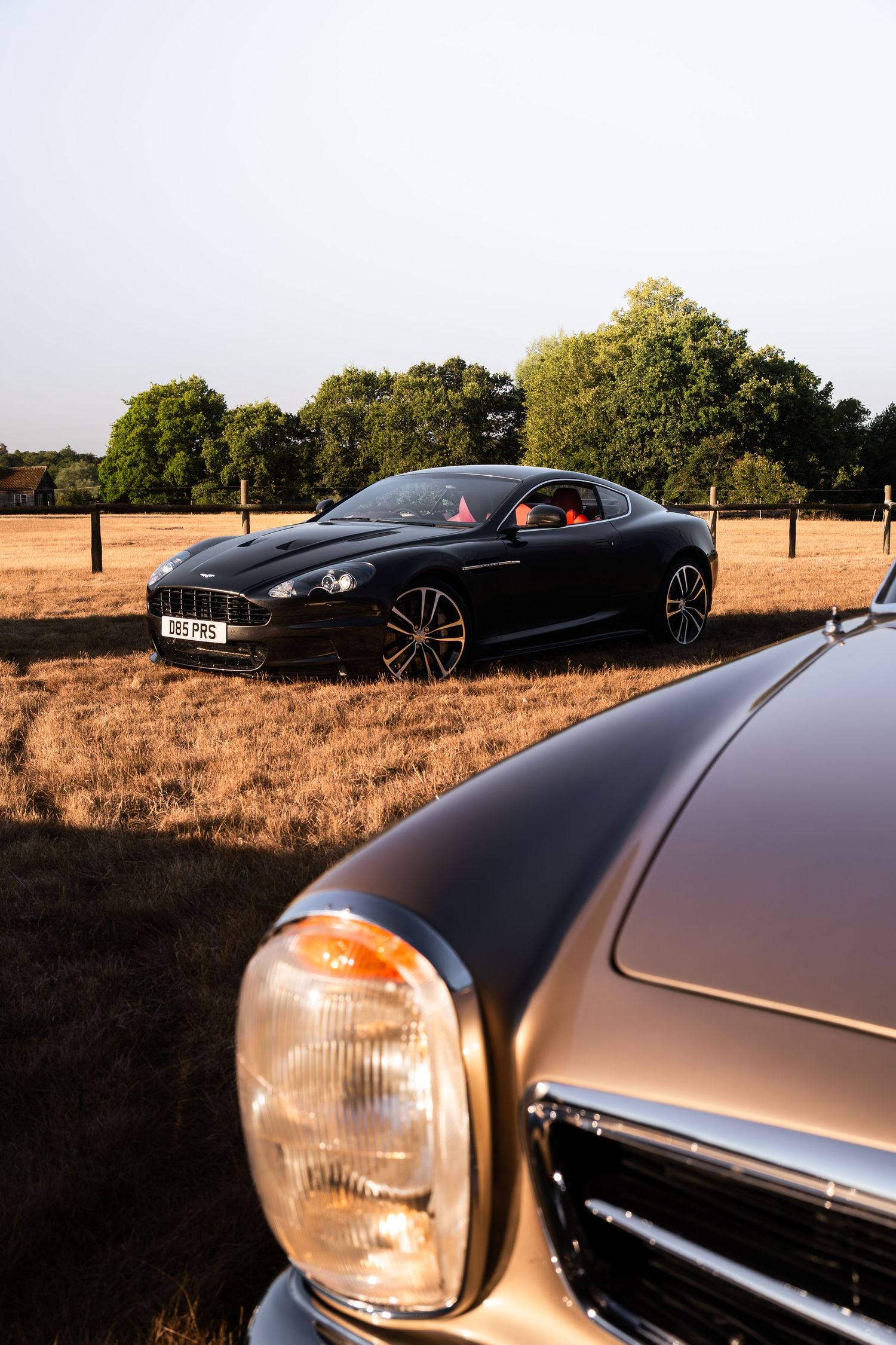 Black Aston Martin DBS coupe parked in golden field with vintage car headlight in foreground.