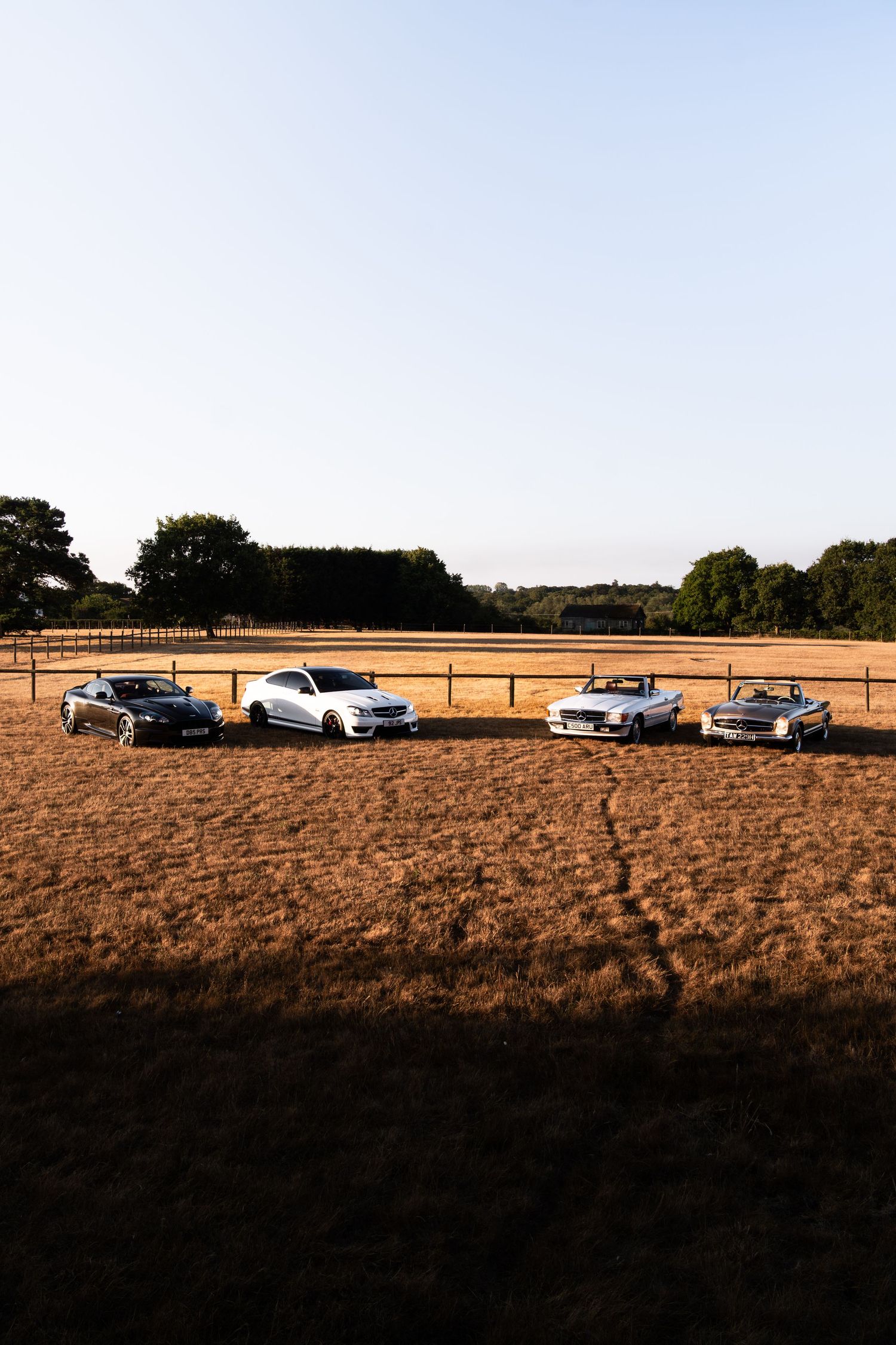 Four luxury cars parked in a golden field at dusk with trees and a wooden fence in the background.