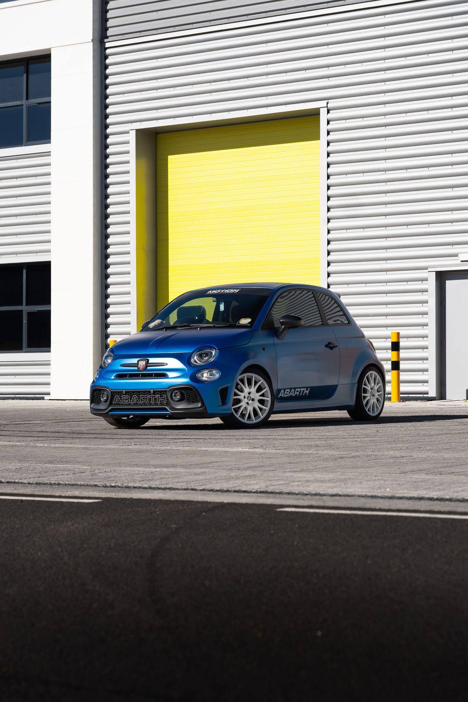 Modified blue Abarth 595 parked outside industrial unit with bright yellow roller shutter door.