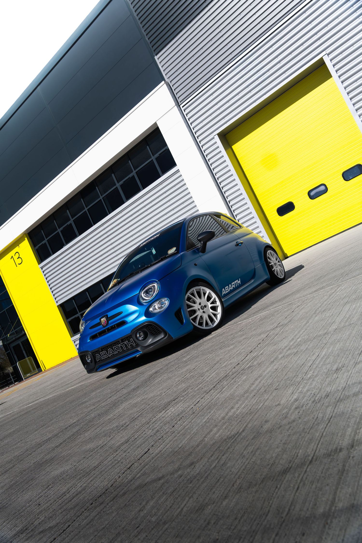 Low-angle shot of a blue Abarth 500 parked outside yellow garage doors at a race track.