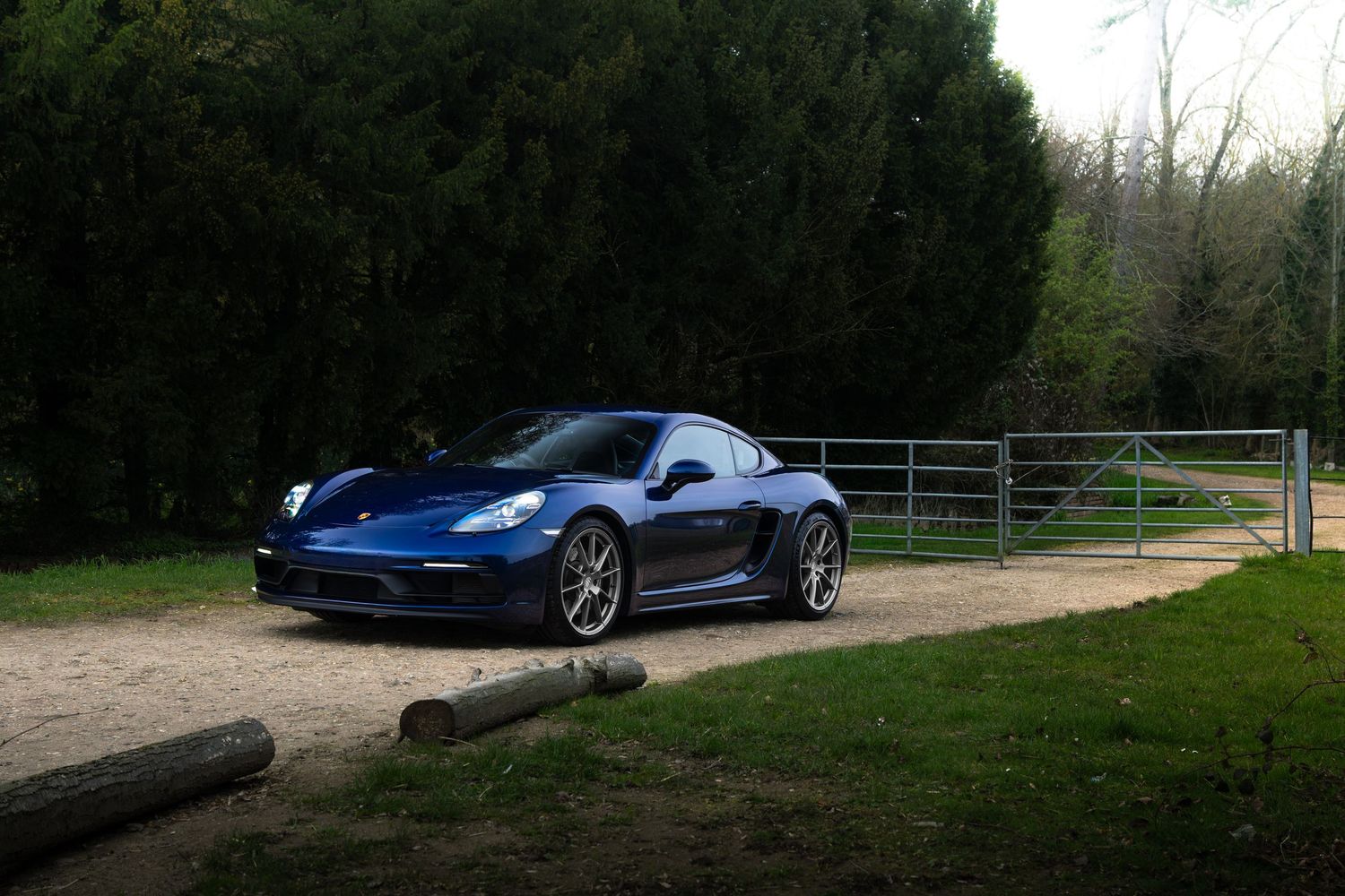 Blue Porsche 718 Cayman GTS parked on gravel driveway beside wooden logs and metal gate surrounded by trees.