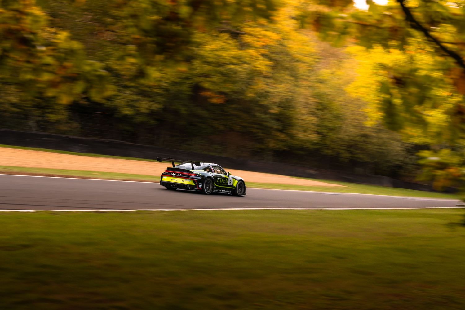 Dark racing car with yellow accents panning shot on circuit surrounded by golden autumn foliage.