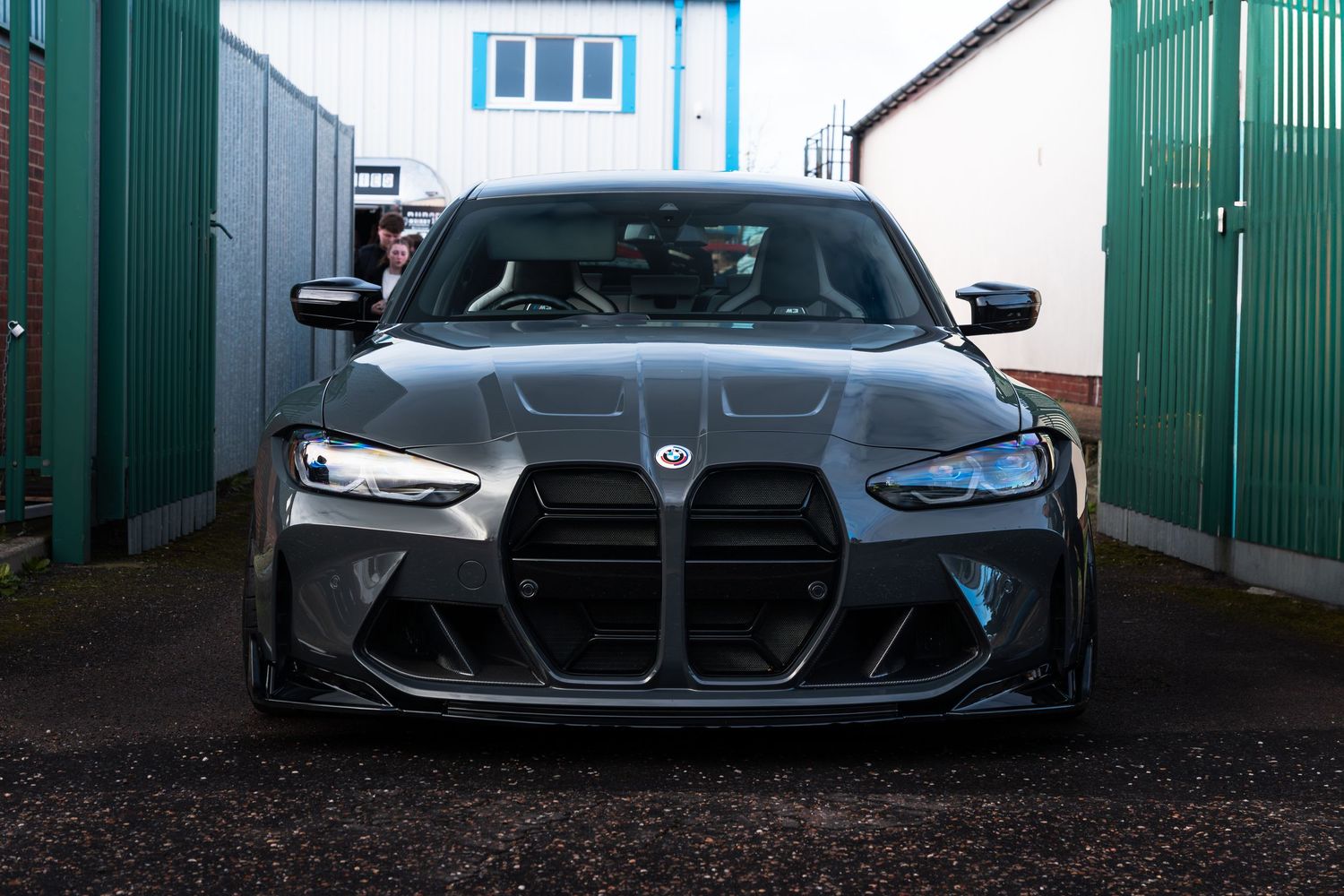 Front view of a dark grey BMW M3 G80 with carbon fiber splitter parked between garage doors.