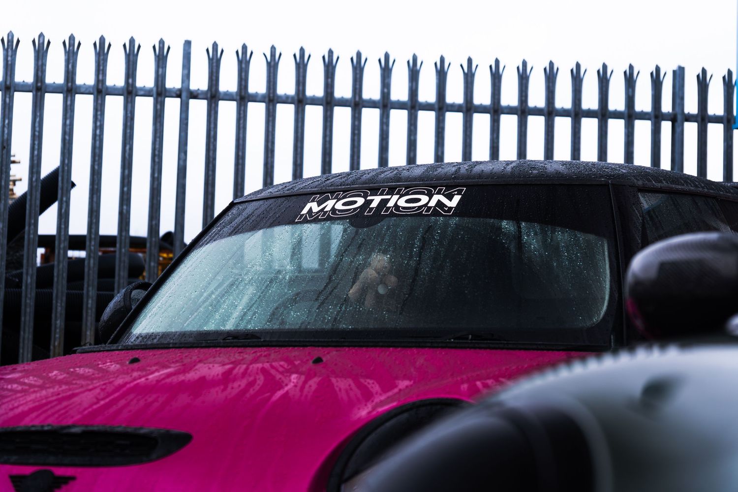 A pink car with Motion Automotive windscreen banner parked behind metal railings.