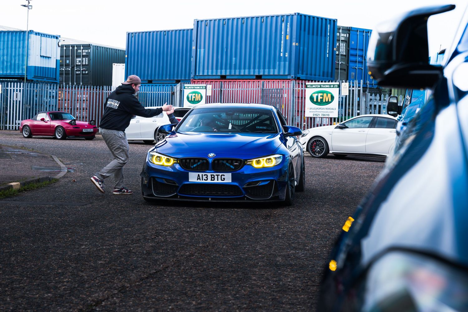 Blue BMW M4 with yellow headlights parked near TFM shipping containers at a car meet.