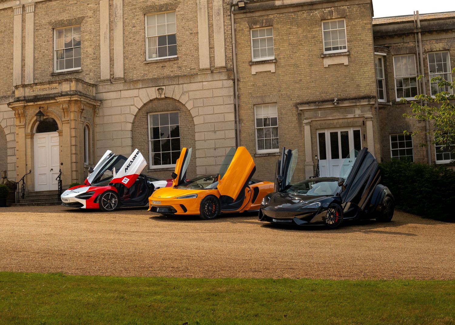 Three McLaren supercars with dihedral doors open parked outside a grand Georgian manor house.