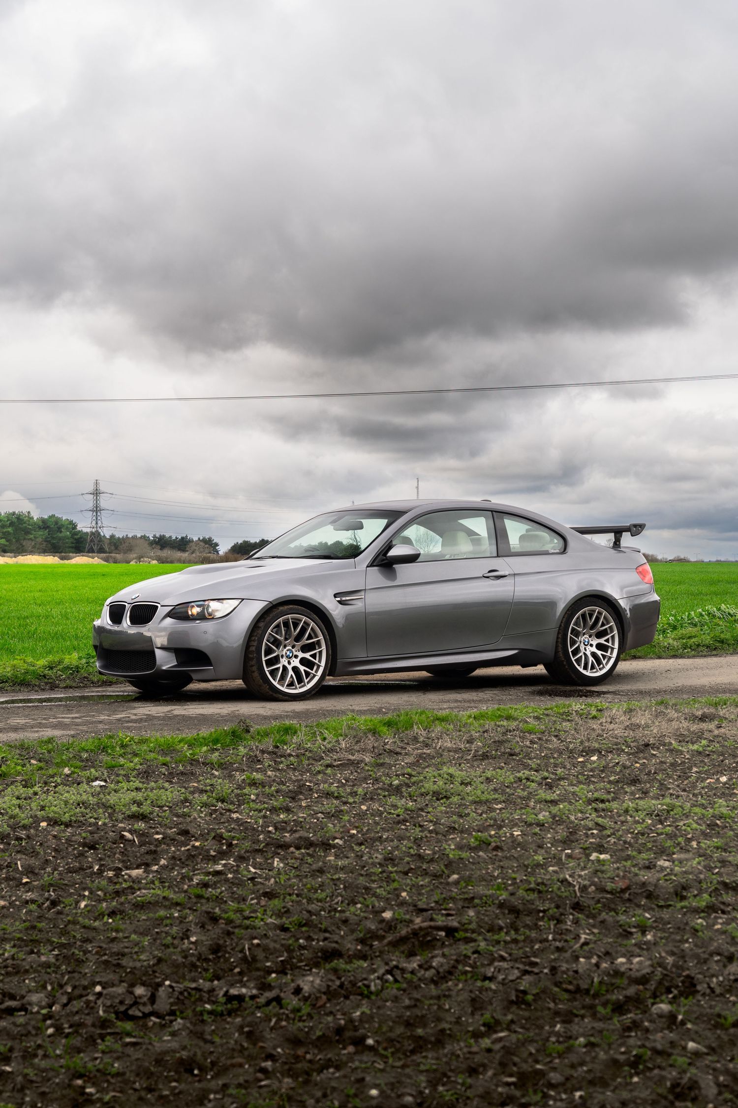 Silver BMW M3 E92 with rear spoiler parked on a country road under dramatic storm clouds.