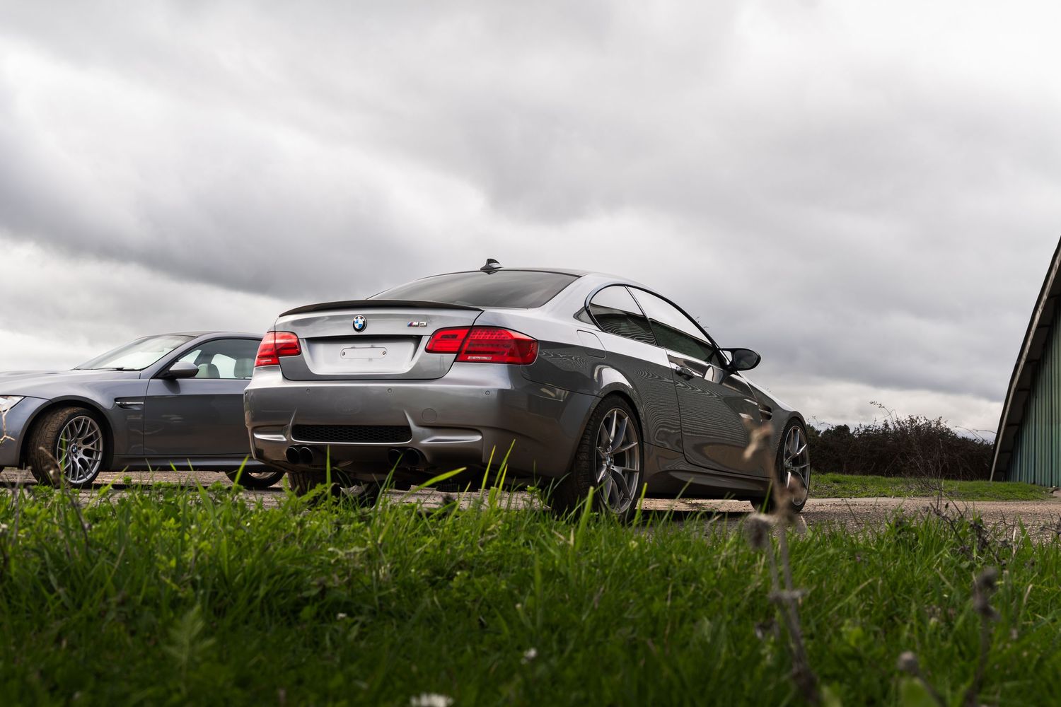 Two gray BMW M3 coupes parked on grass with a cloudy sky and green field background.