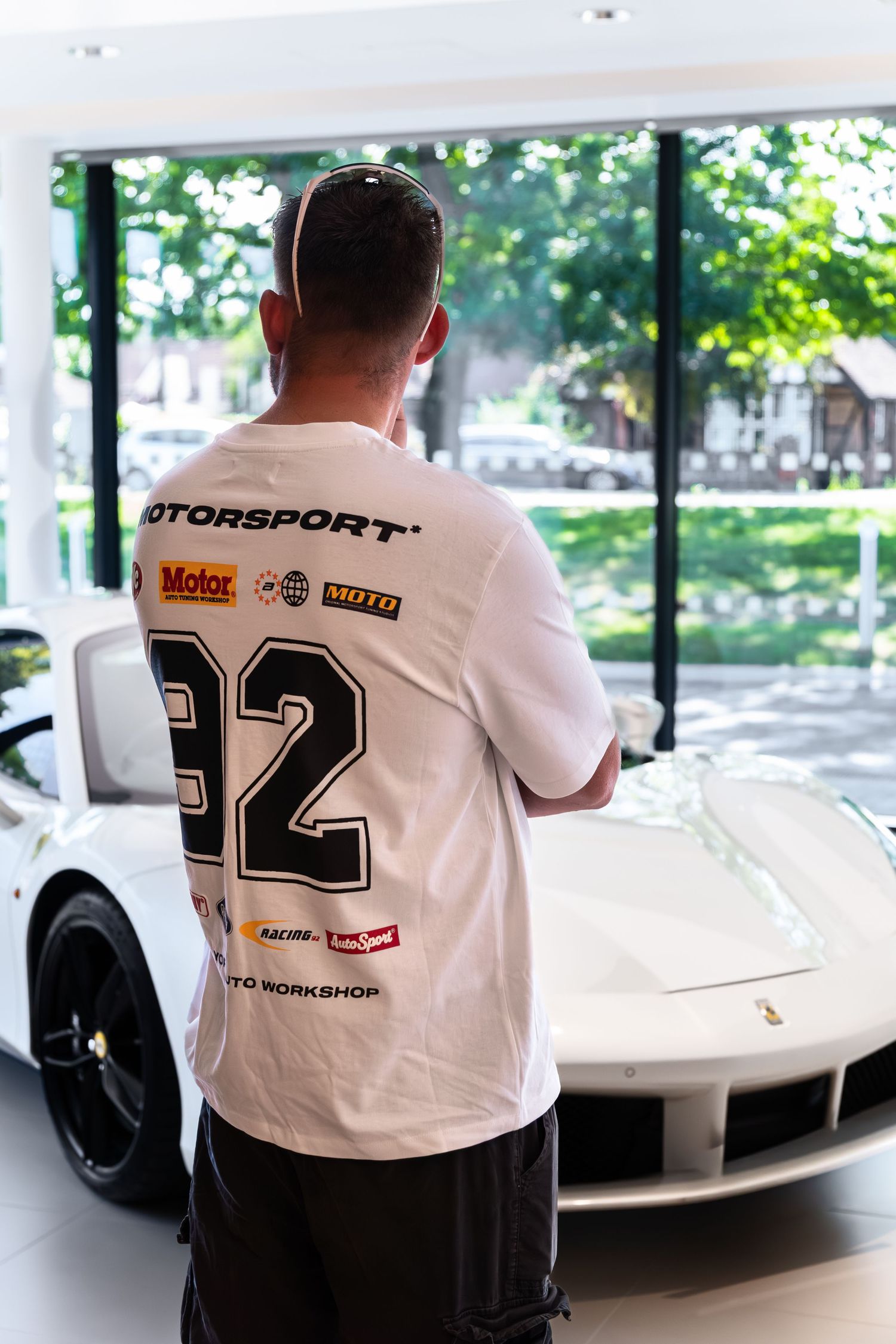 Man in white Motorsport number 32 t-shirt admiring a white Ferrari in a luxury car showroom.