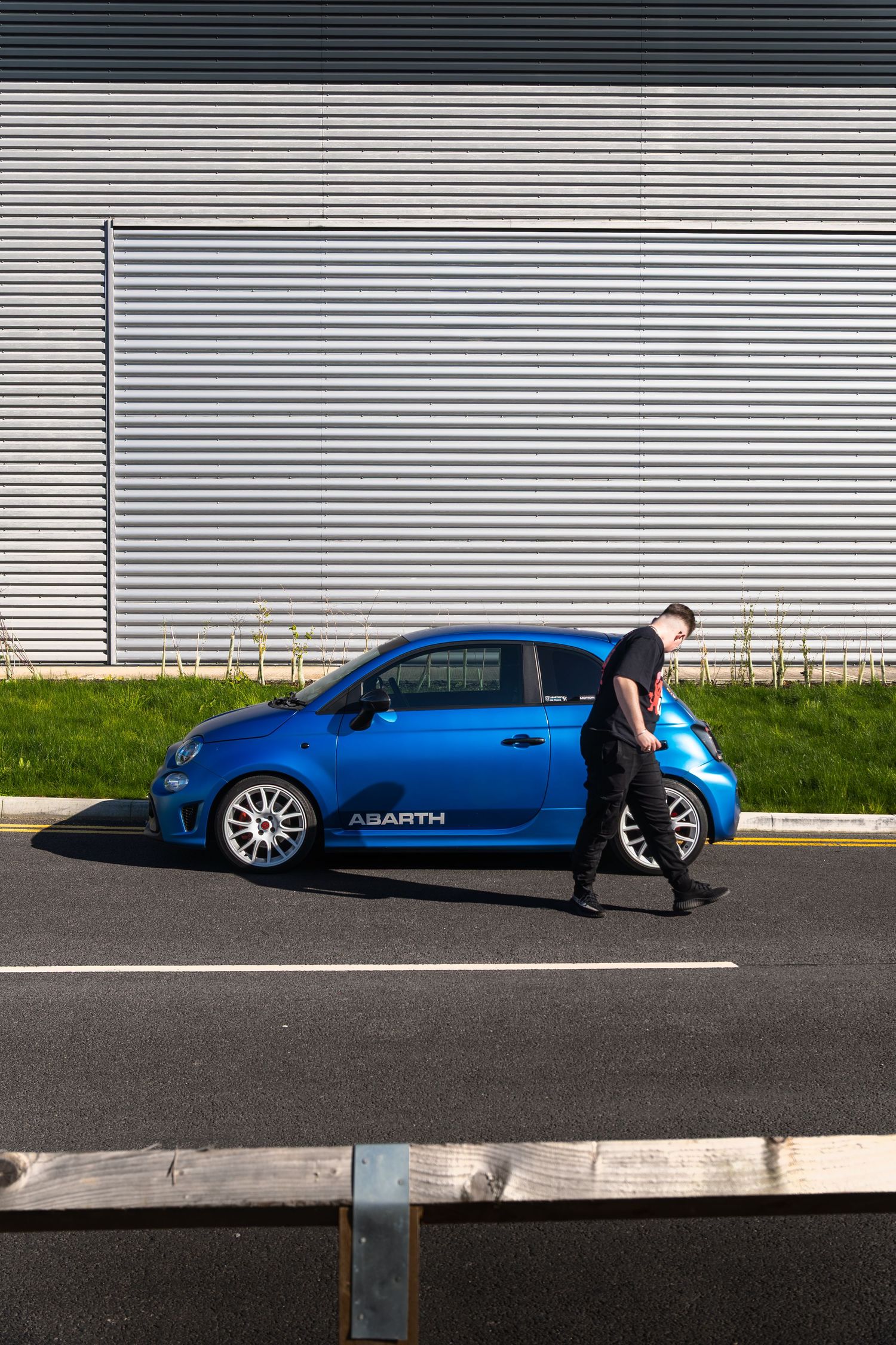 Man in black t-shirt walking past a blue Abarth 500 parked beside a corrugated metal warehouse wall.