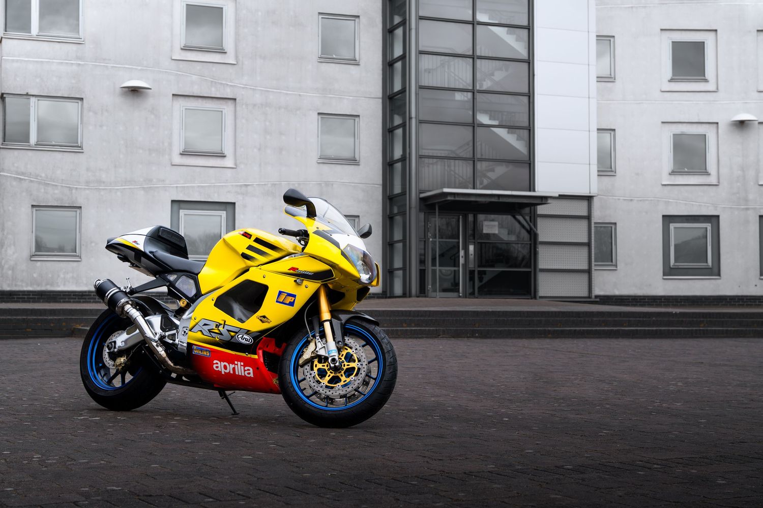 Yellow Aprilia RSV-R superbike parked in front of a modern building, showcasing its sleek fairing and sporty design.