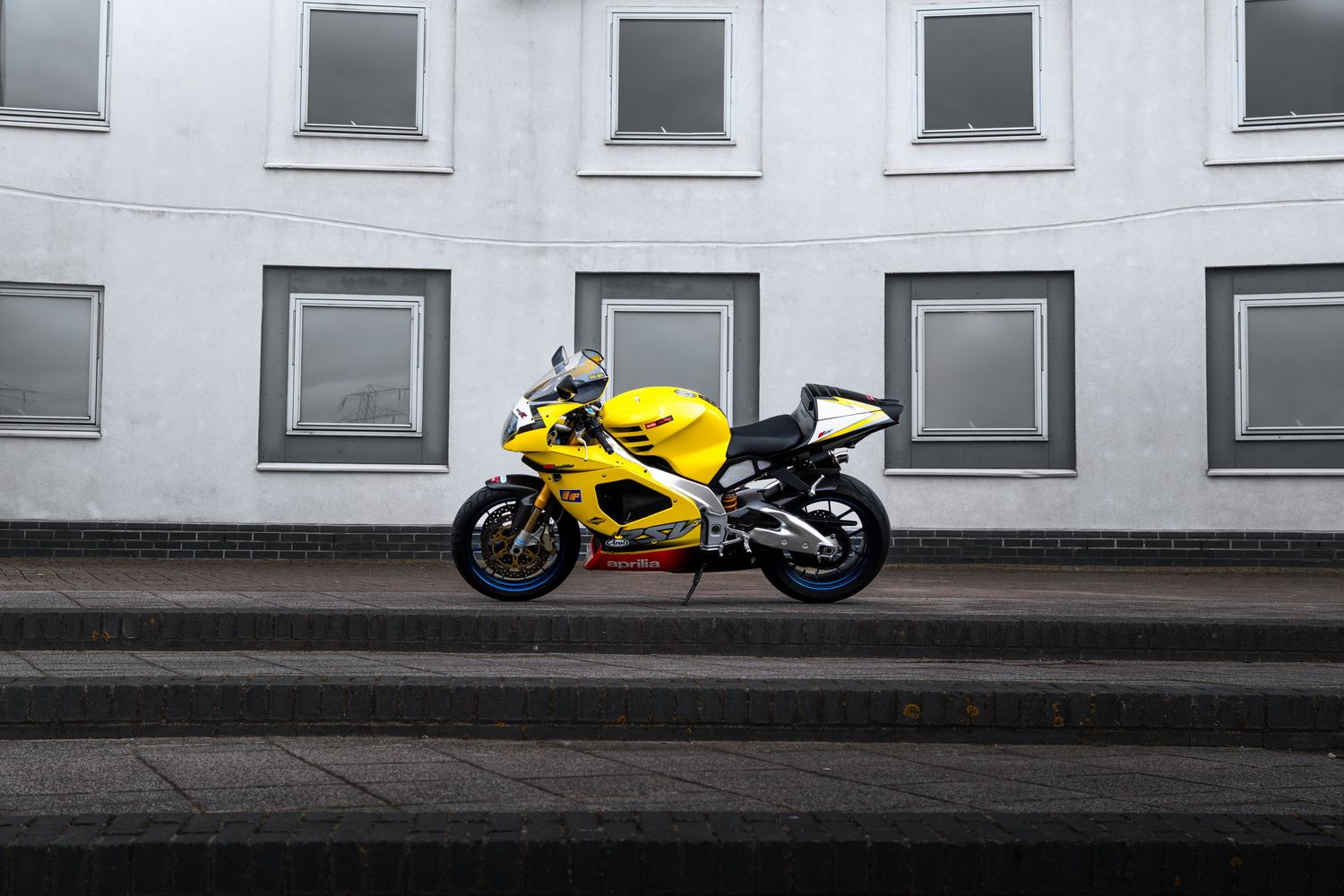 Side profile of yellow Aprilia RSV-R sport motorcycle parked near a curved white building exterior.