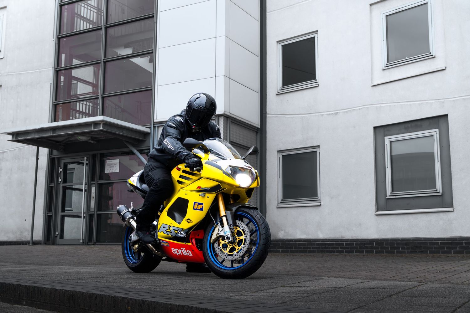 Motorcyclist on bright yellow Aprilia RSV superbike maneuvering in urban setting.