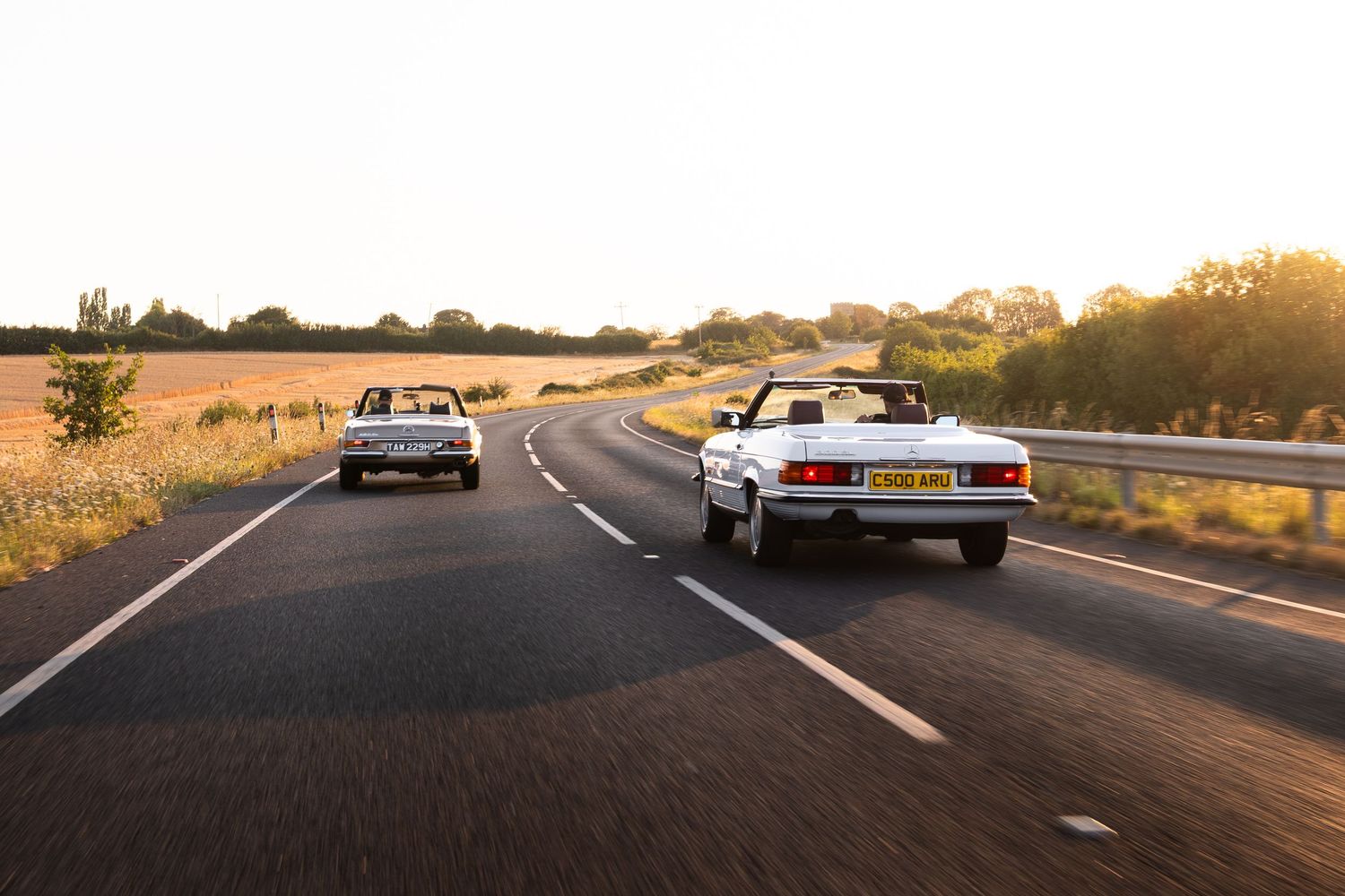 Two classic convertible cars driving on a scenic highway at golden sunset hour with fields in the background.