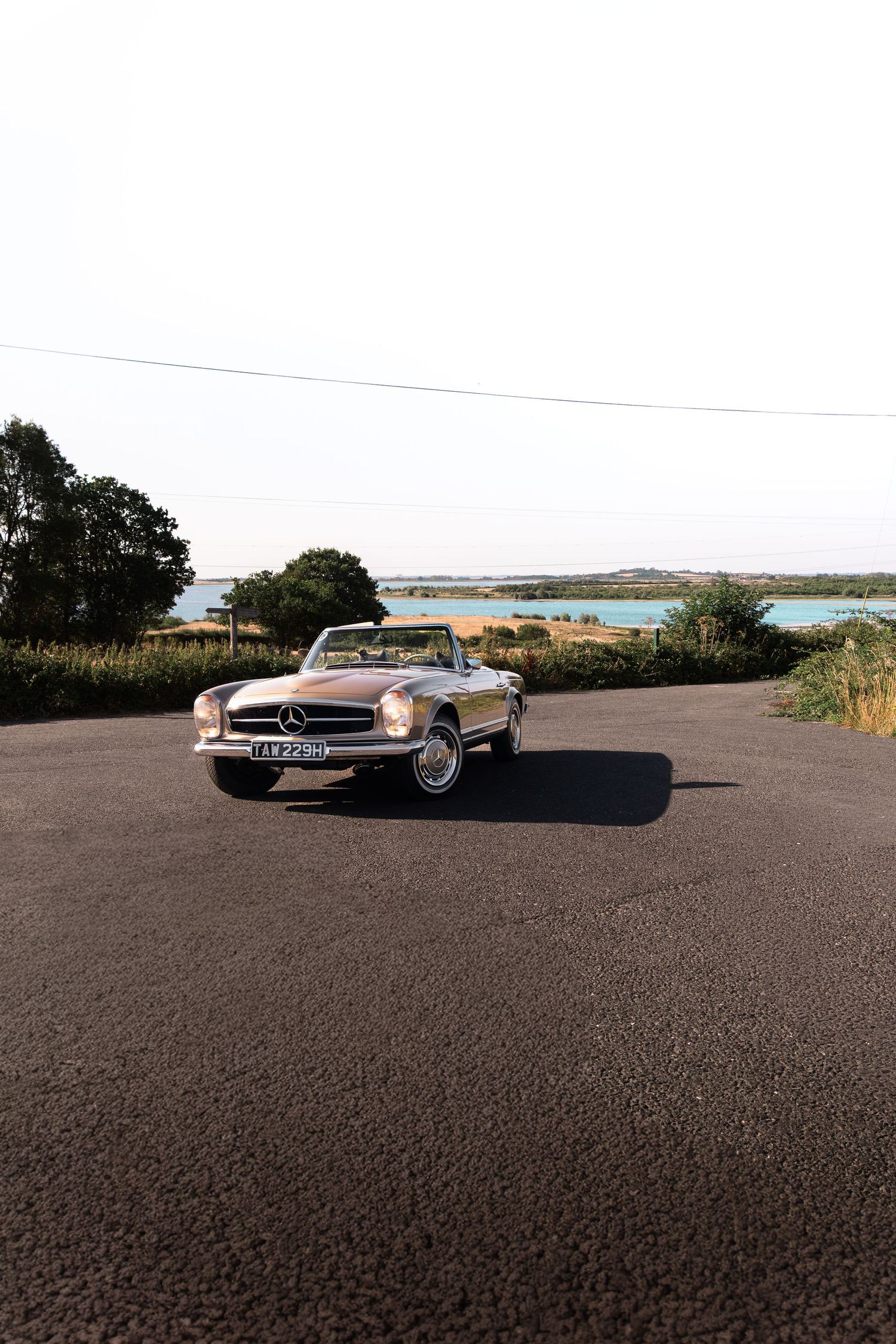 Classic Mercedes-Benz SL convertible parked on a rural road with scenic water views and green trees in background.